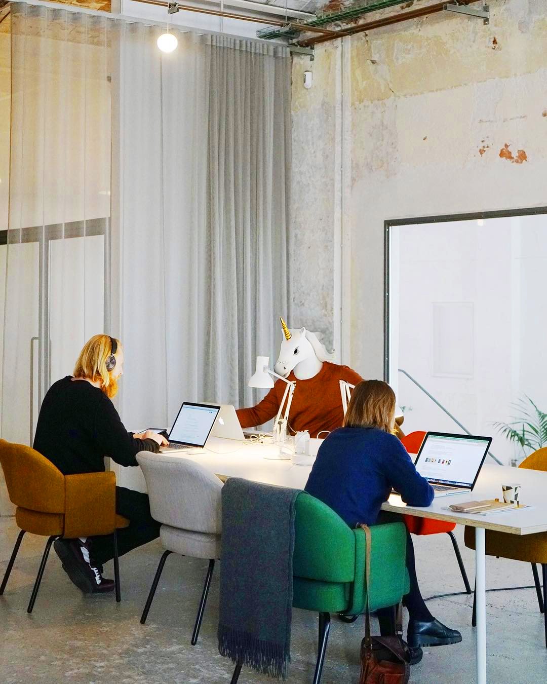 Office workspace with three employees working on laptops at a long white table. One employee wears a horse mask with a unicorn horn, and another has headphones on. The room has a modern, industrial style with exposed pipes and a large window.