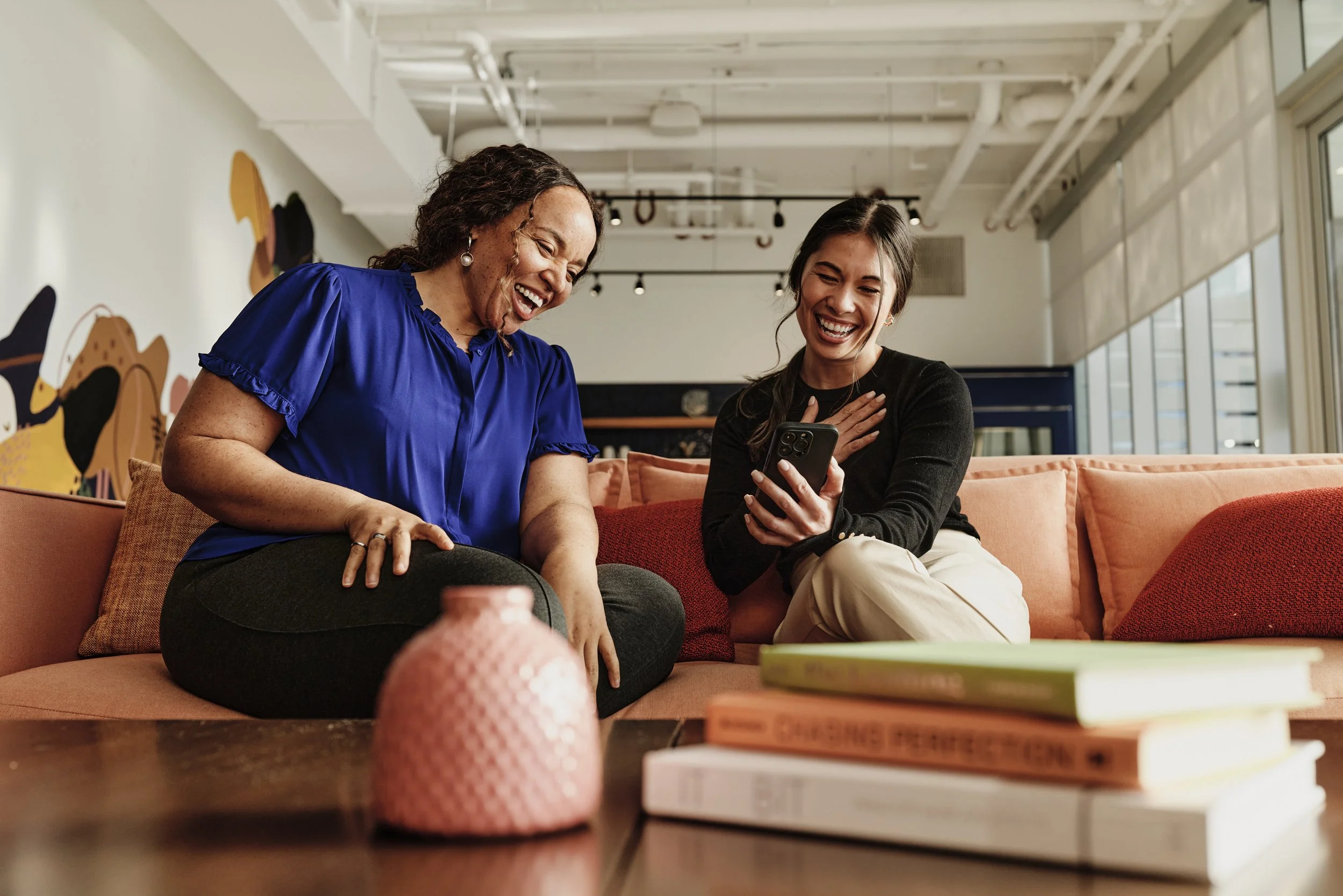 Two women sitting on a peach-colored sofa, laughing and looking at a smartphone. One woman wears a blue top and the other a black top. There are books on the table in front of them and a pink textured vase.