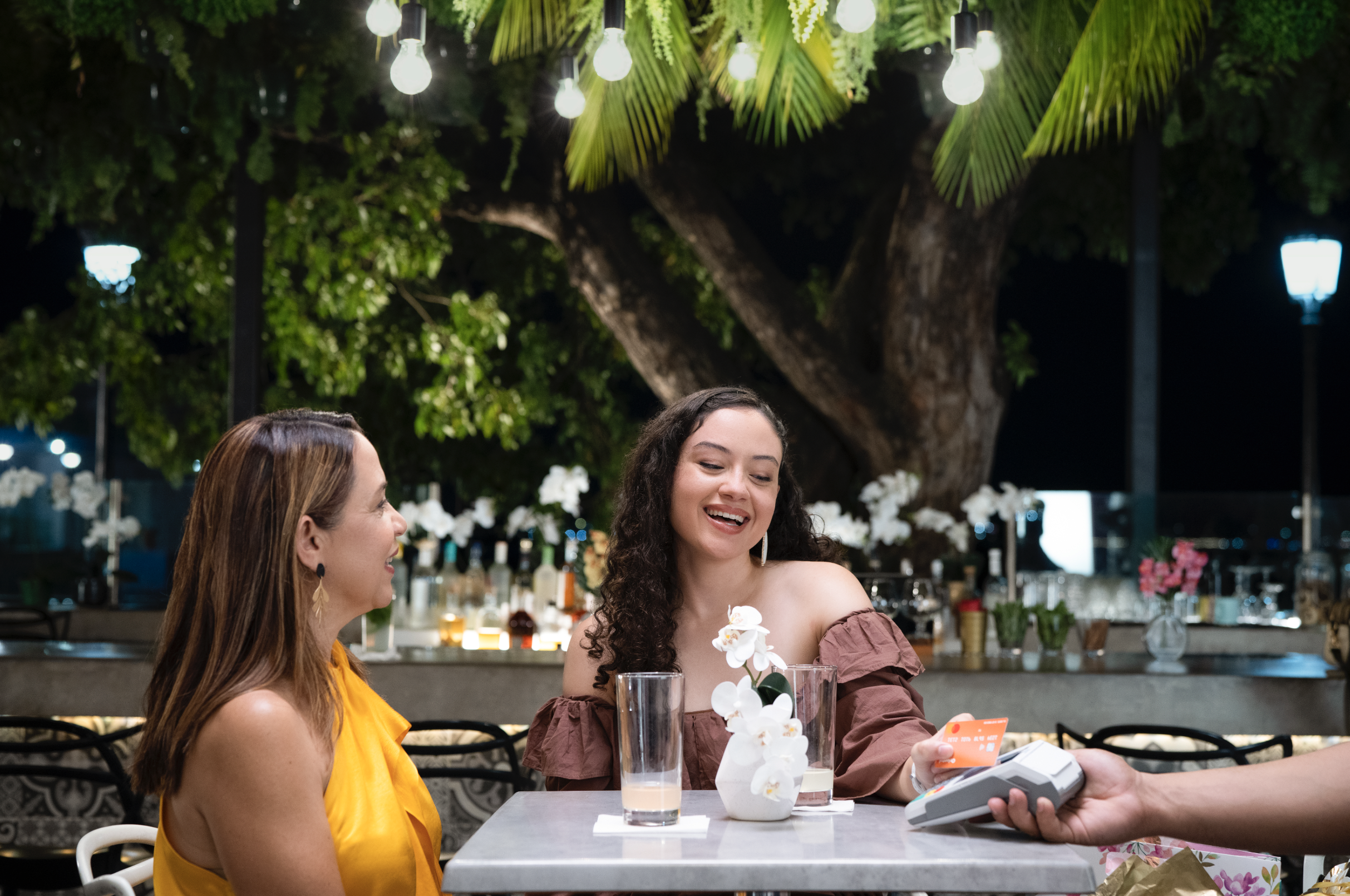 Two women sitting at a table outdoors at night, smiling and paying with a credit card, with a bartender handling a card reader. Behind them, there is a large tree with hanging lights and a bar with bottles and flowers.