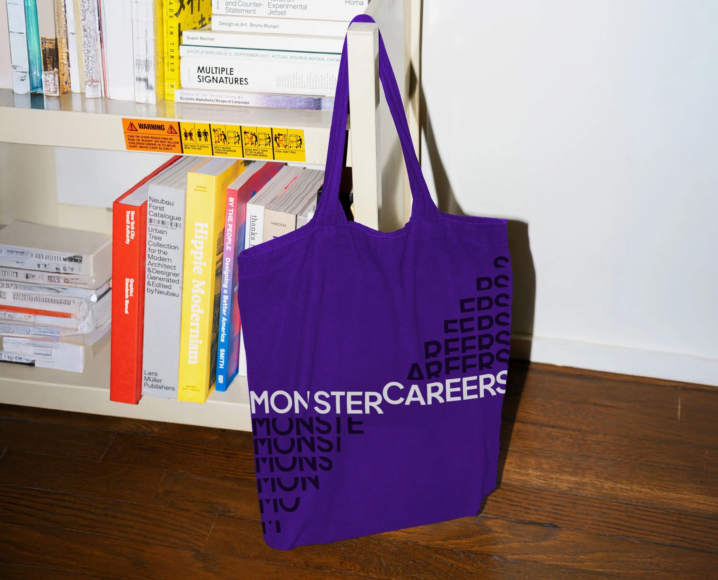 A purple tote bag with black and white text hanging on a white shelf filled with books, placed on a wooden floor.