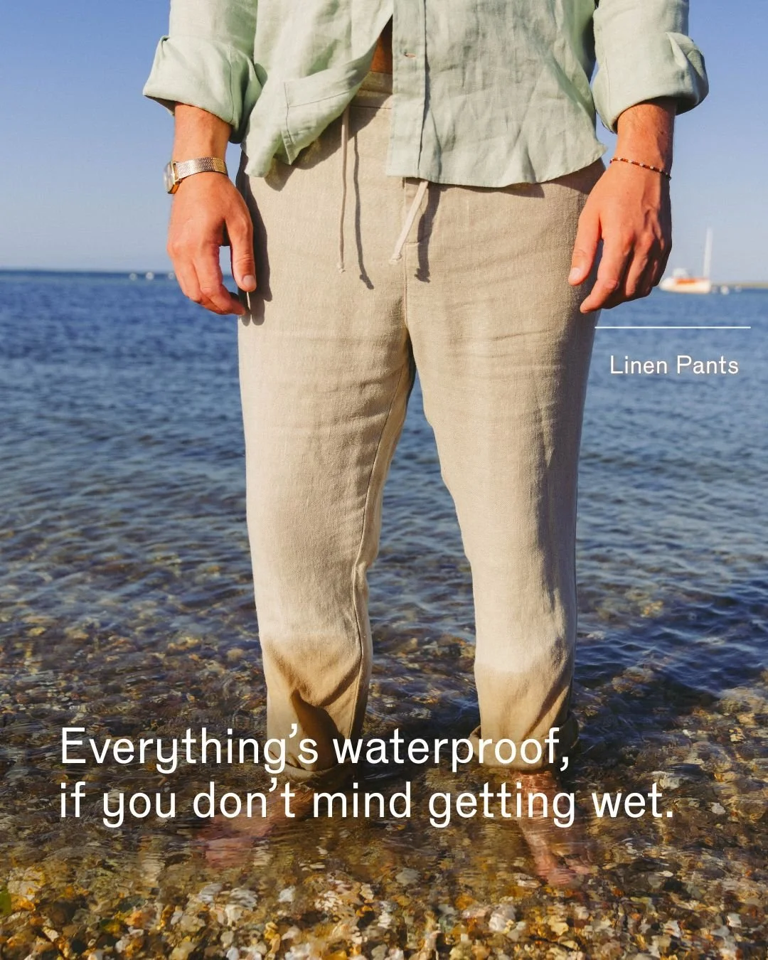 Person standing in shallow water wearing beige linen pants and a light green shirt, with the ocean and a boat in the background.