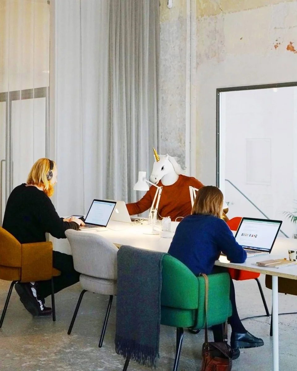 People working in an office with a person wearing a unicorn mask seated at the table.