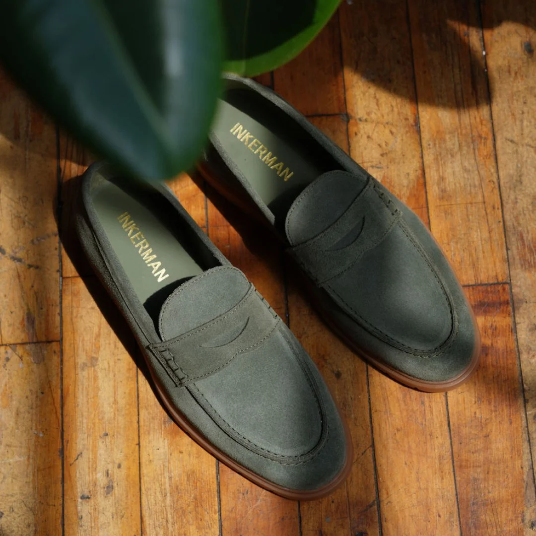 Pair of grey leather loafer shoes placed on a wooden floor, with part of a green plant in the upper left corner.