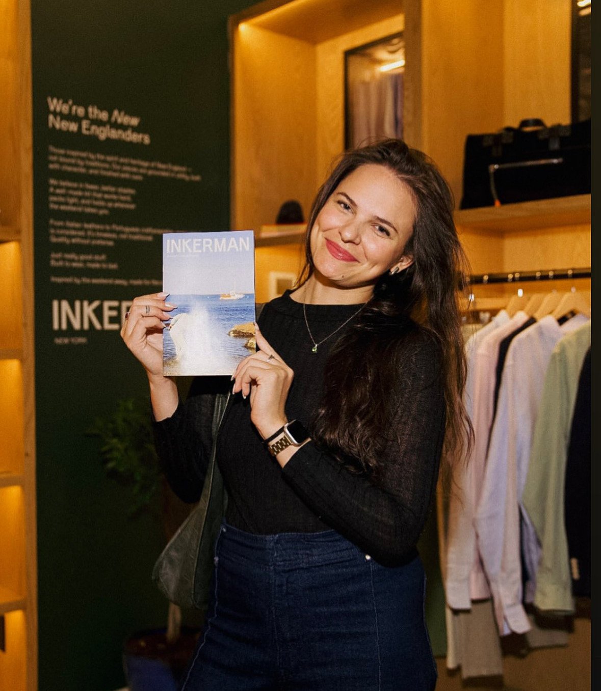 A woman with long dark hair wearing a black sweater, smiling, holding up a book titled "INKERMAN" with a beach scene on the cover, inside a clothing store or boutique.