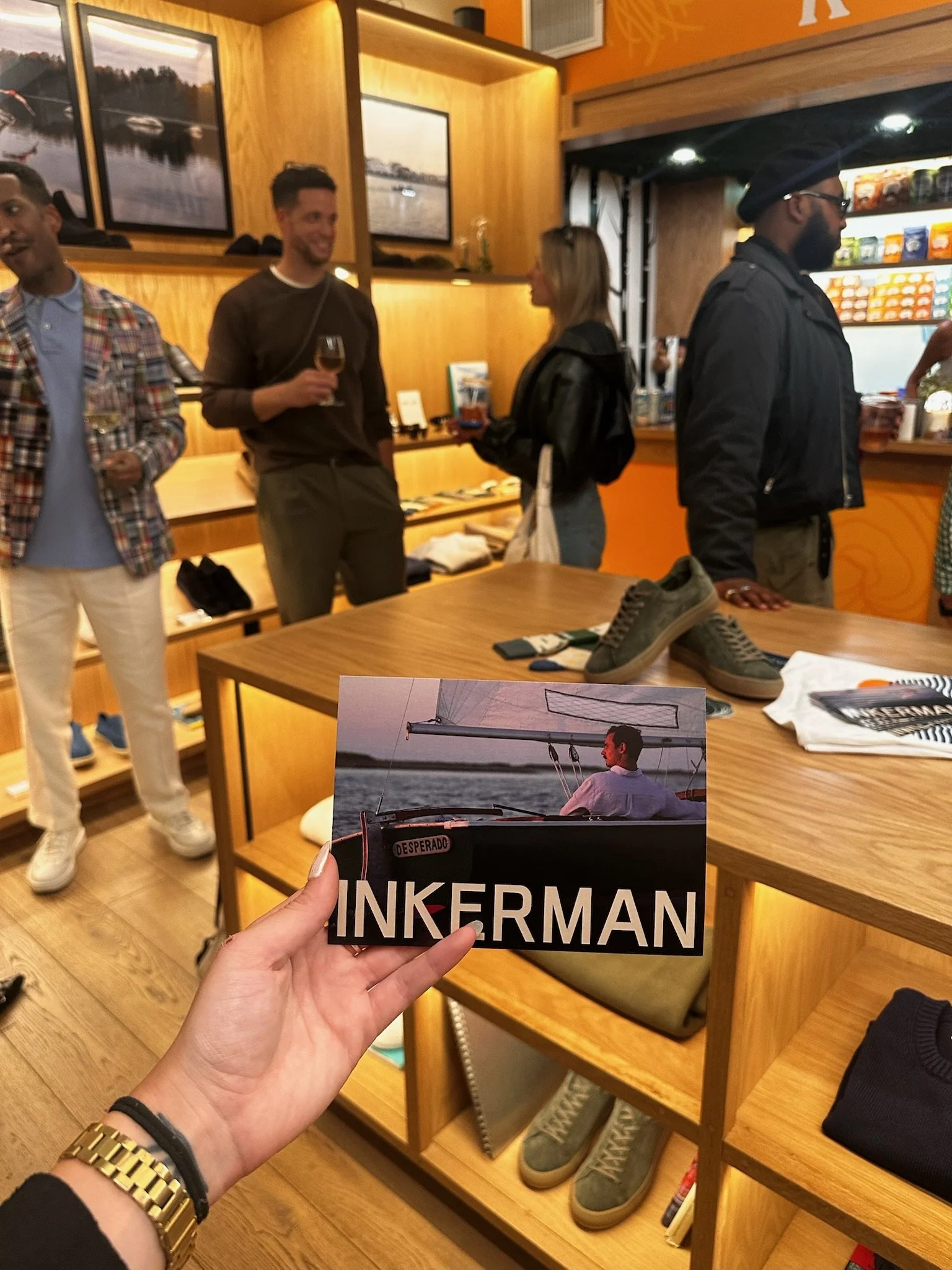 Inner view of a retail store with several people shopping and talking. Featured in the foreground is a hand holding a magazine with the title 'INKERMAN' and an image of a man sailing on the cover.