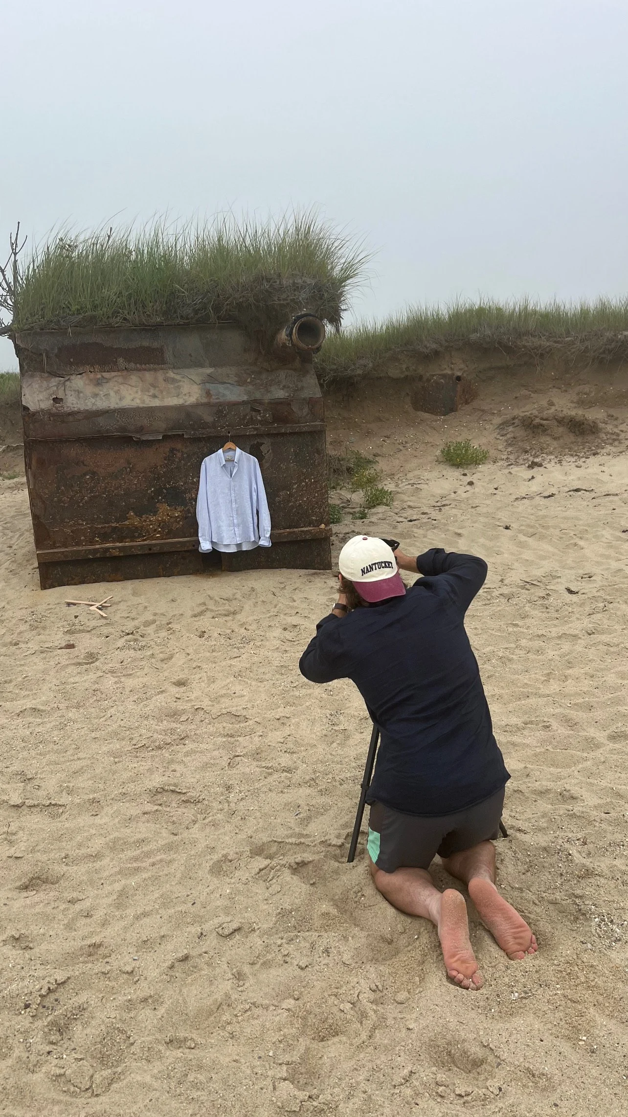 A person kneeling on sand taking a photograph of a shirt hanging on a rusty, makeshift outdoor display against a cloudy sky.