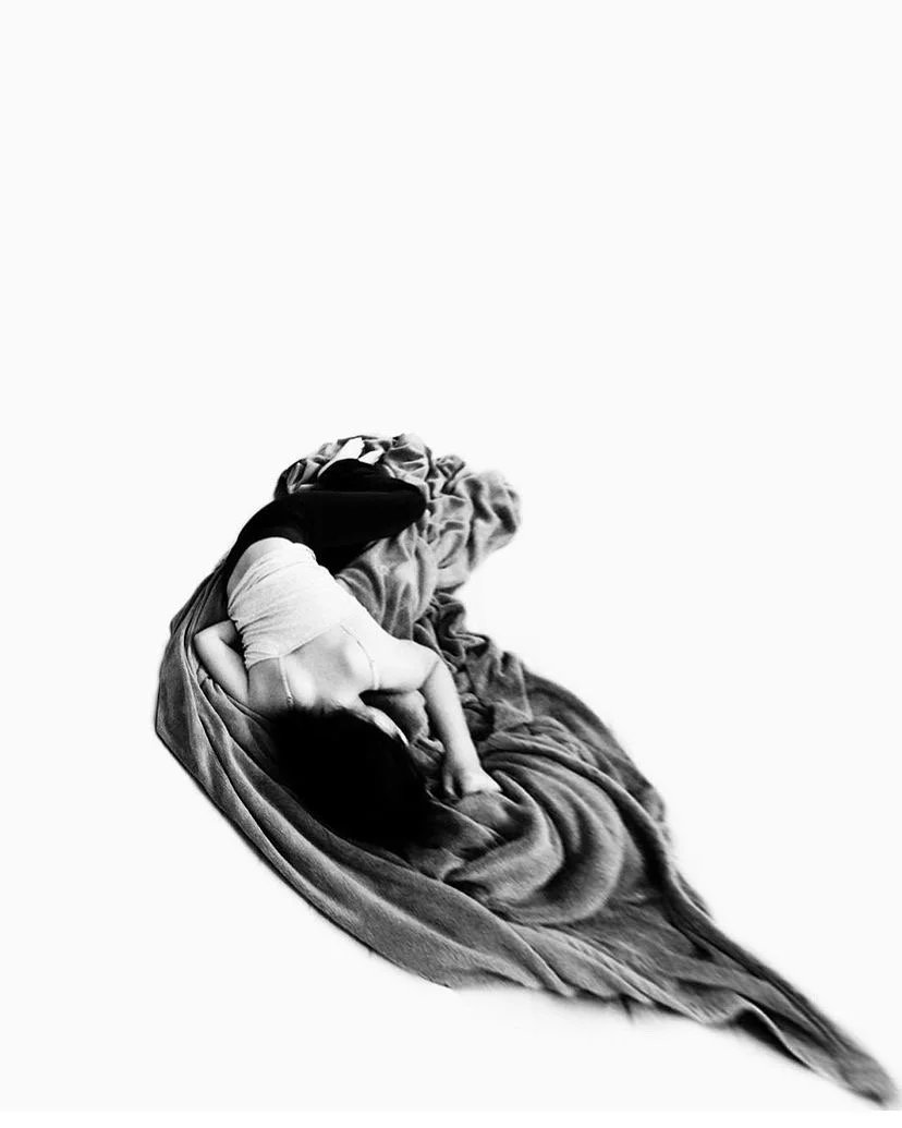 Black and white photo of a person lying on a large, soft blanket, curled up in a fetal position, with hair covering their face.