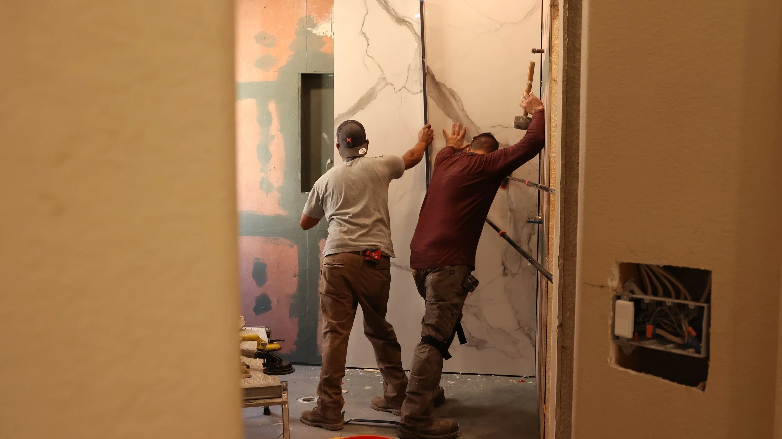 Two workers are installing a large marble or stone wall panel, working together to position it accurately, in a room under construction.