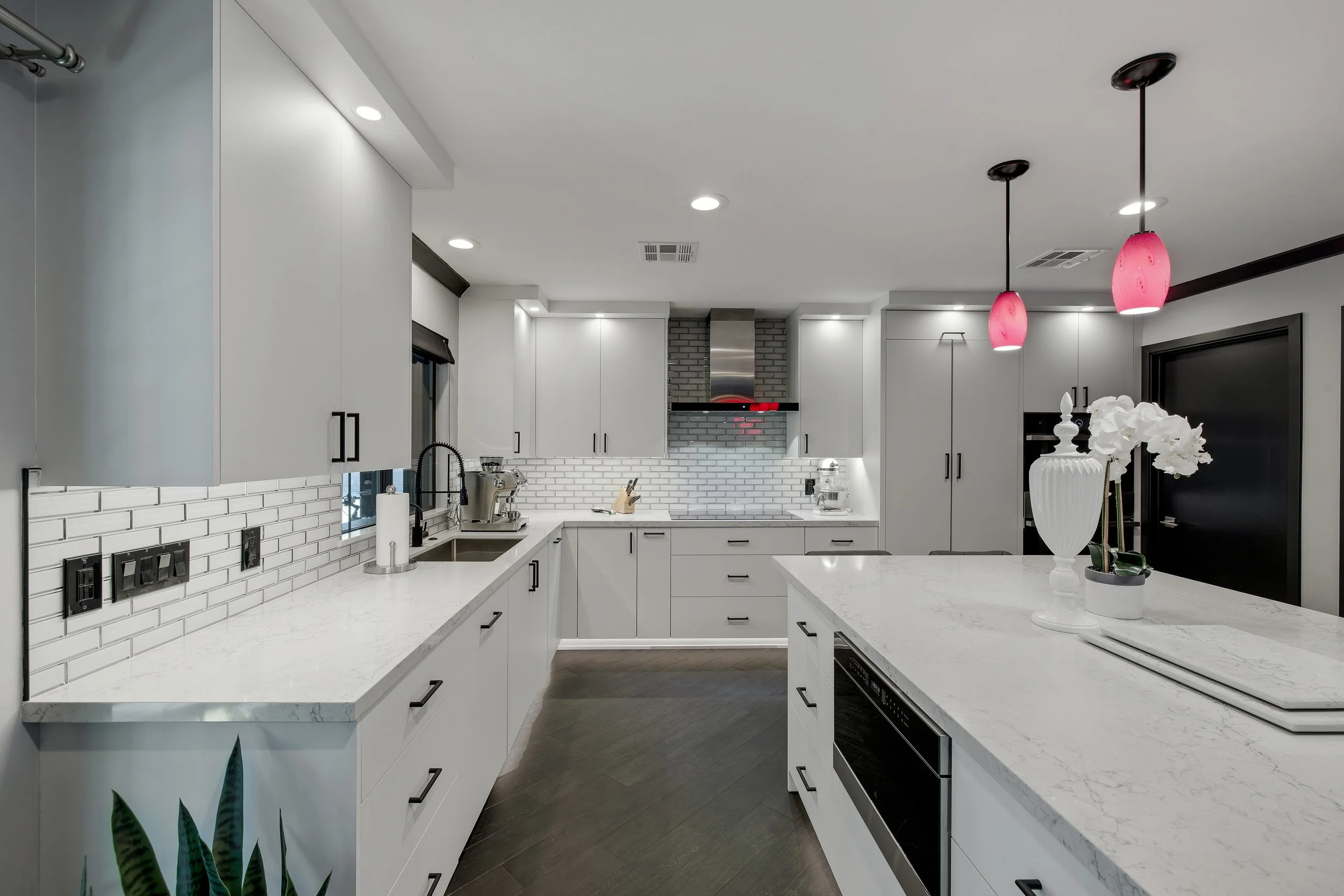 Modern kitchen with white cabinetry, marble countertops, black handles, white subway tile backsplash, stainless steel appliances, black window frame, pink pendant lights, white vase with flowers, and dark wood flooring.