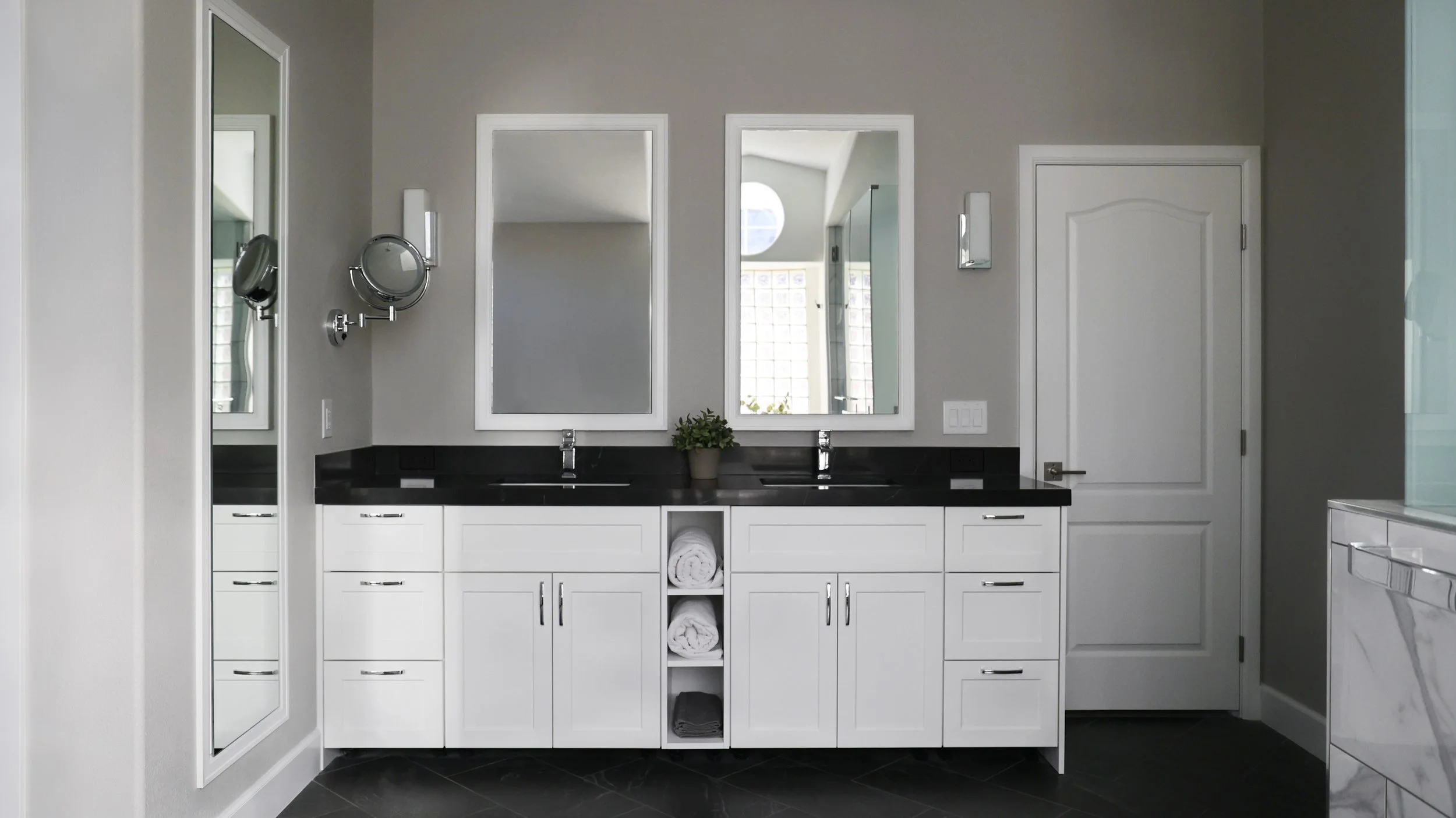 Modern bathroom with white cabinets, dual sinks, black countertops, and large mirrors, with a small potted plant between the mirrors.