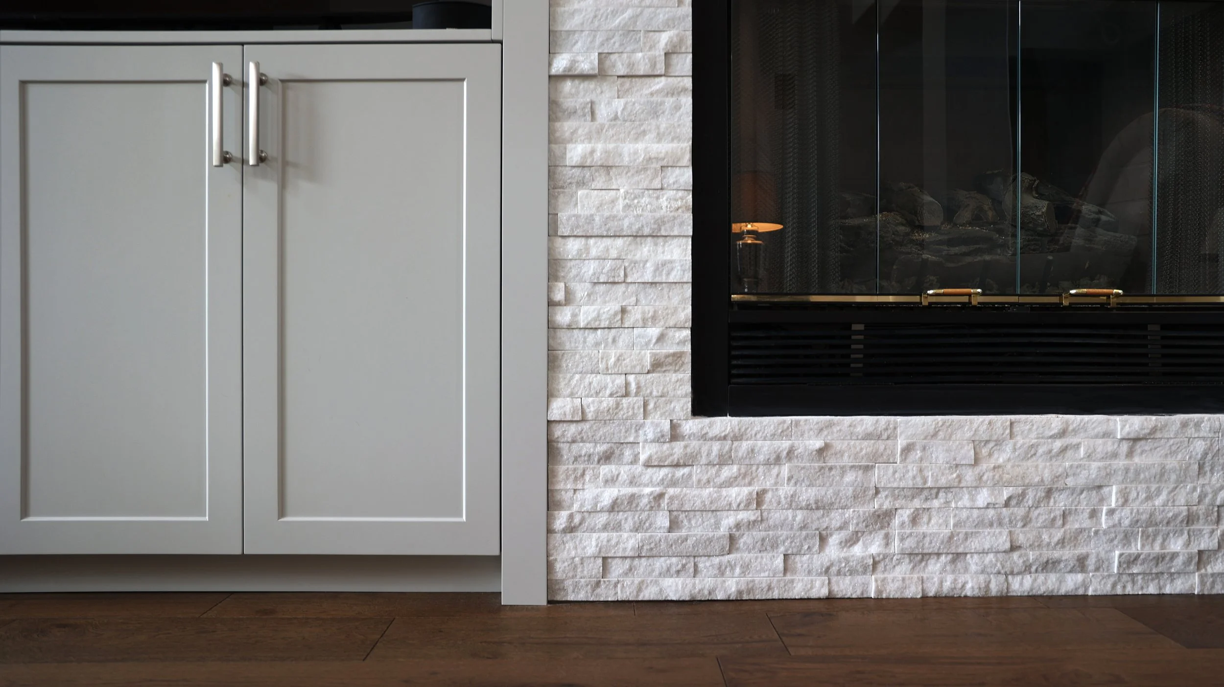 Close-up of a modern white kitchen cabinet with brushed metal handles next to a stone-textured wall and a black fireplace with glass doors.