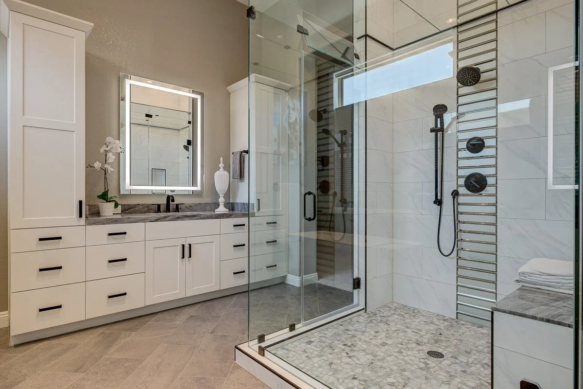 Modern bathroom with a glass-enclosed shower, white cabinetry with black handles, a large backlit mirror, a gray countertop, and a neutral-colored tiled floor.