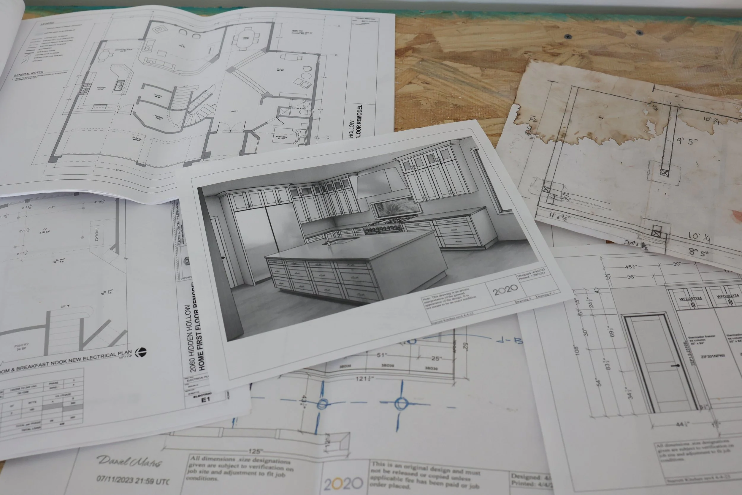 Blueprints and architectural plans spread out on a wooden surface, including detailed drawings of an interior kitchen and house layout, with some sketches on aged paper.
