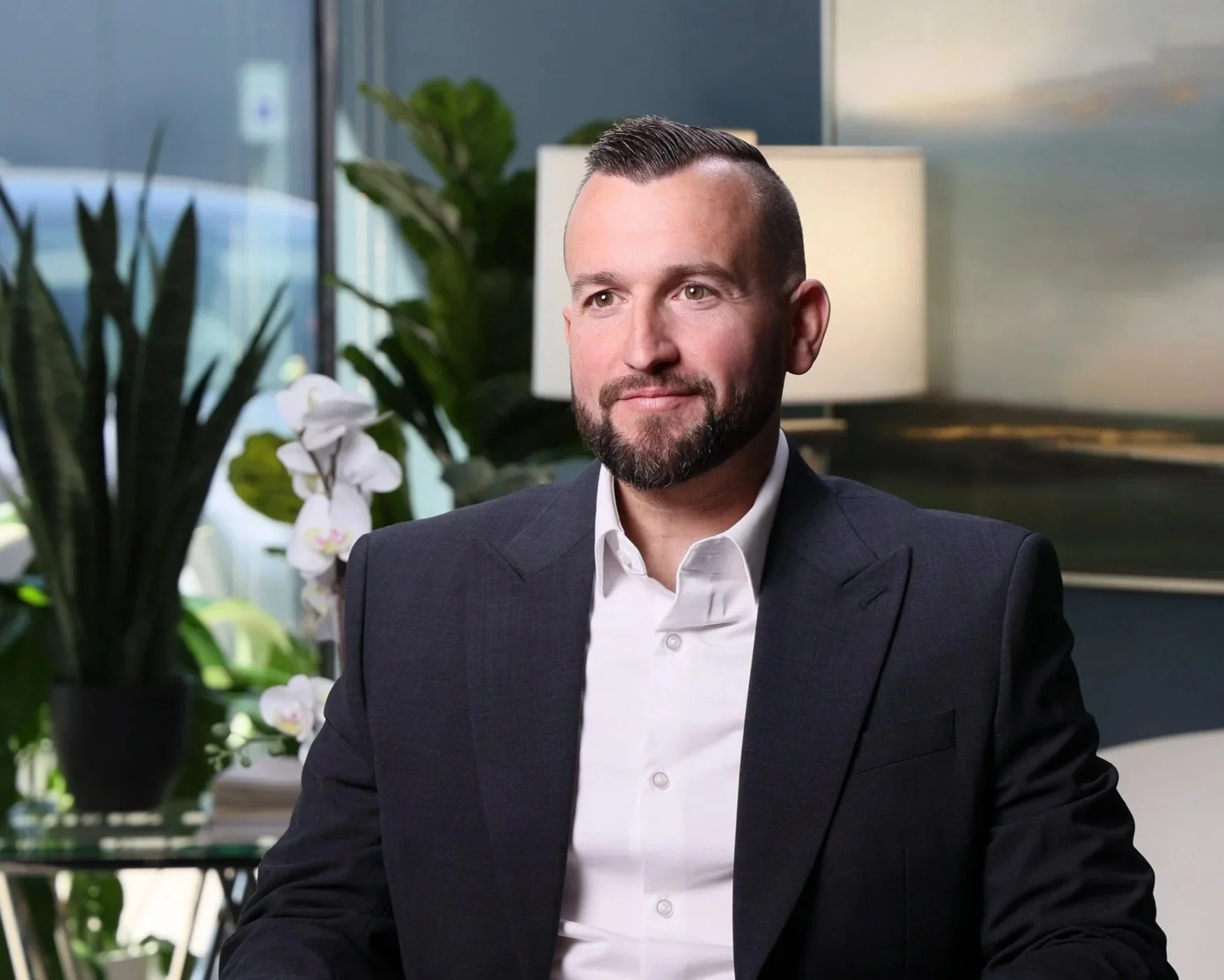 A man with a beard, dressed in a dark suit and white shirt, sitting in an office with plants and a window in the background.