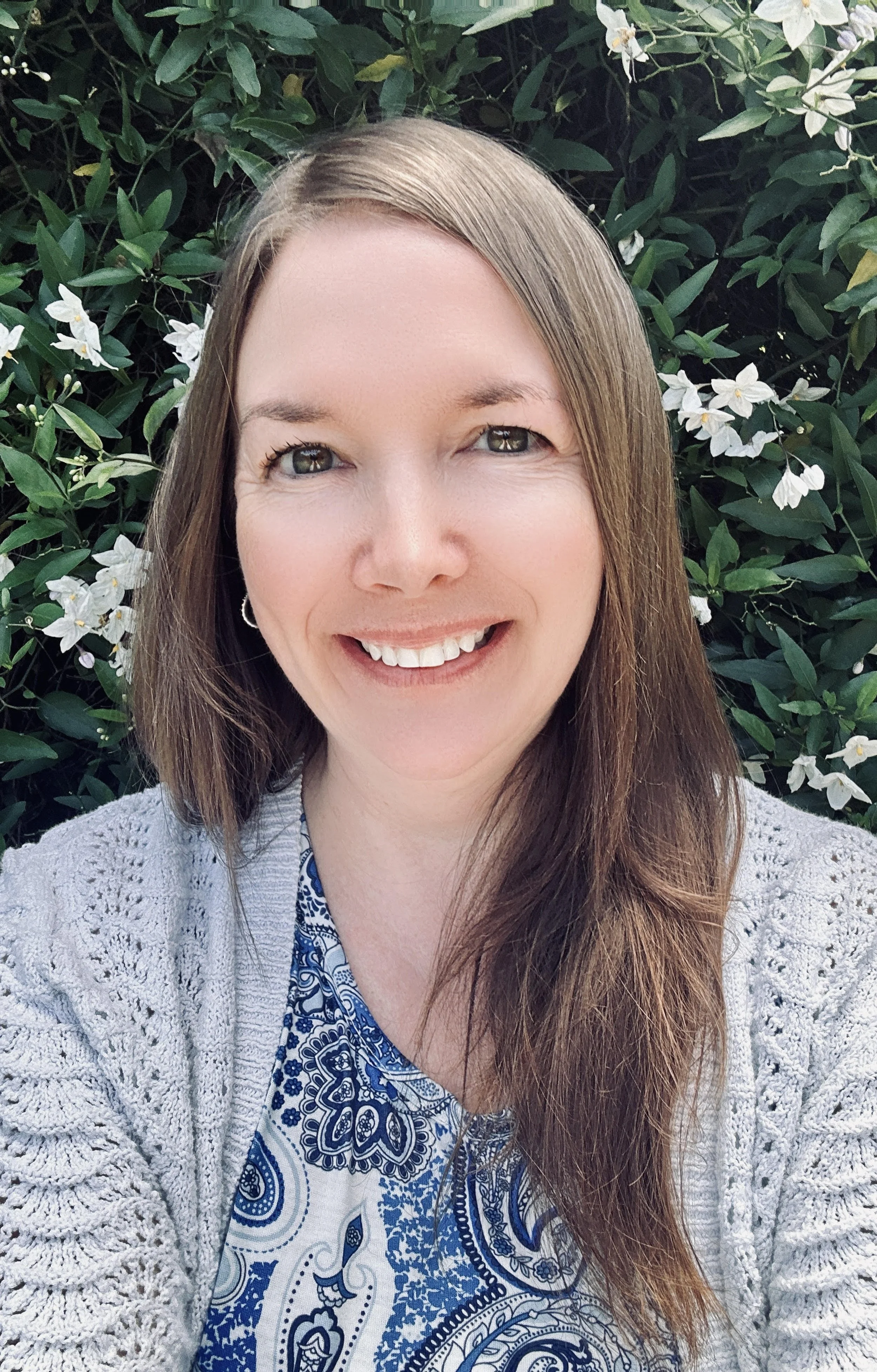 A smiling woman with long brown hair, wearing a patterned blue and white shirt and a gray knitted sweater, standing in front of green leafy bushes with white flowers.