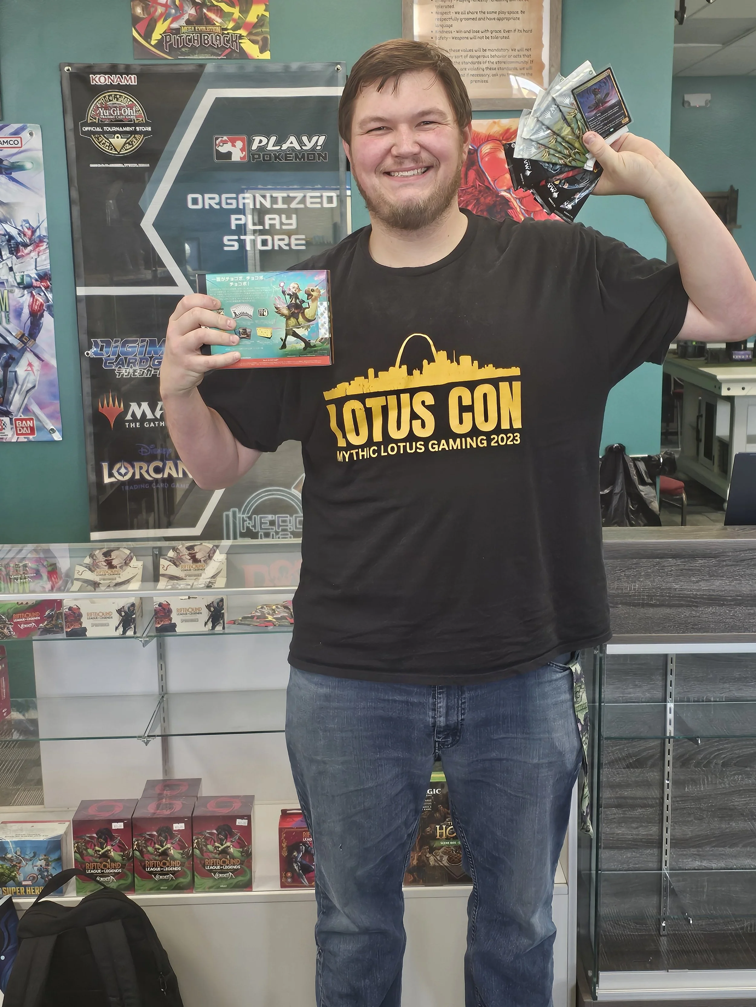 A smiling man holding trading cards and a small box in a game store with shelves of board games and card packs behind him, and various posters featuring trading card games and Pokémon on the wall.