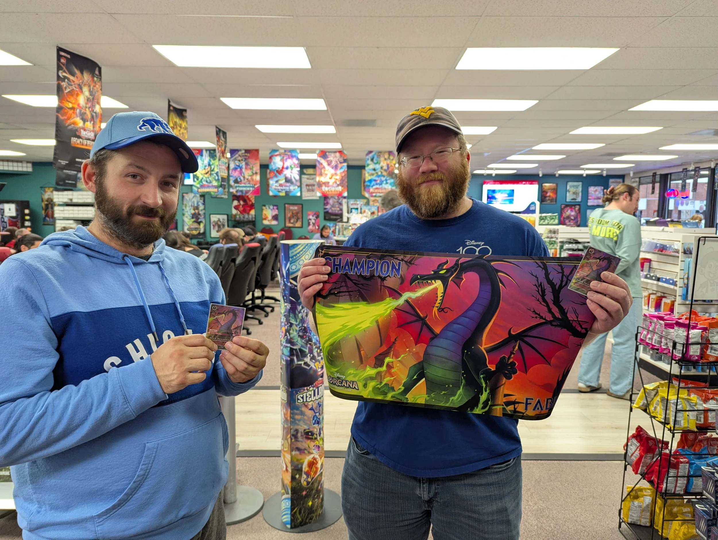 Two men standing in a store, holding large Pokémon trading card game booster packs and smaller cards. The store has posters of Pokémon and other card games on the walls, with other shoppers in the background.