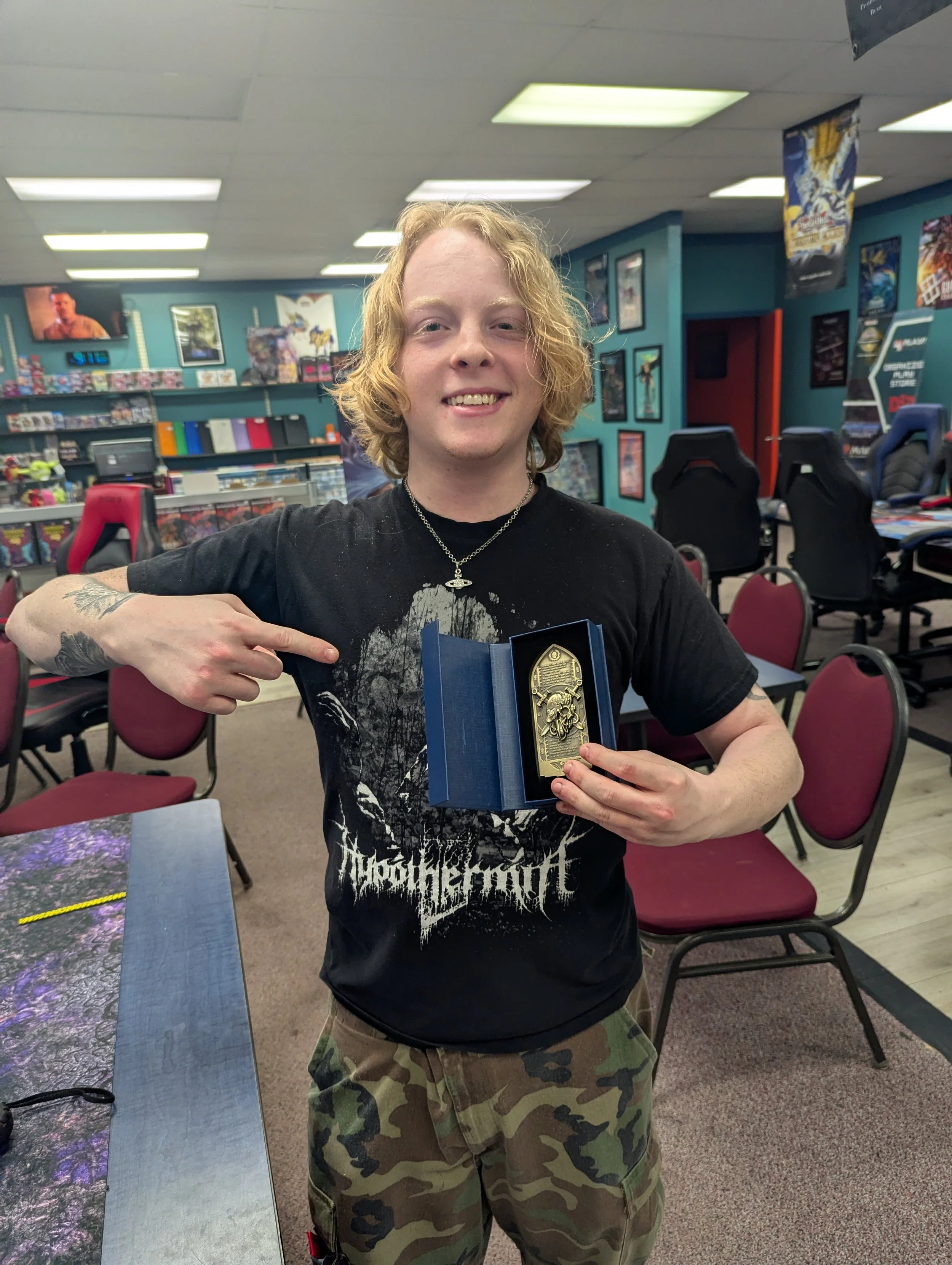 A young man with long, curly red hair smiling and pointing at a medal in a blue box. He is wearing a black T-shirt with white graphics, a silver necklace, and camouflage pants inside a gaming or hobby store.