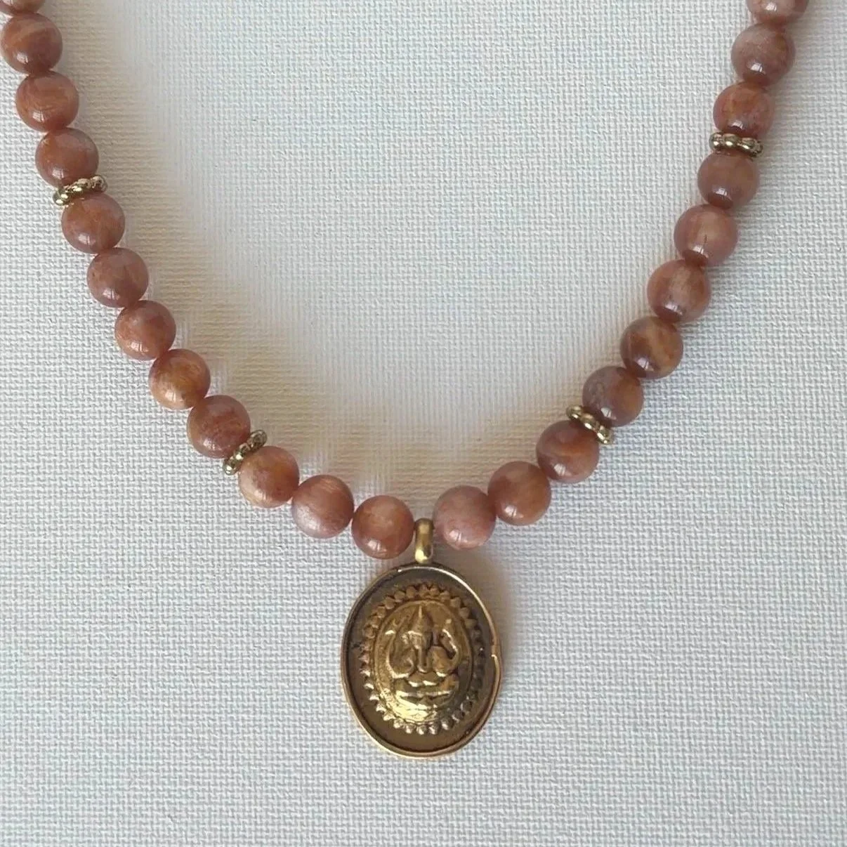 A unique handmade beaded necklace with pinkish-brown Sunstone gemstone beads and small gold spacers, featuring an oval gold pendant with an engraved face of Lord Ganesha.