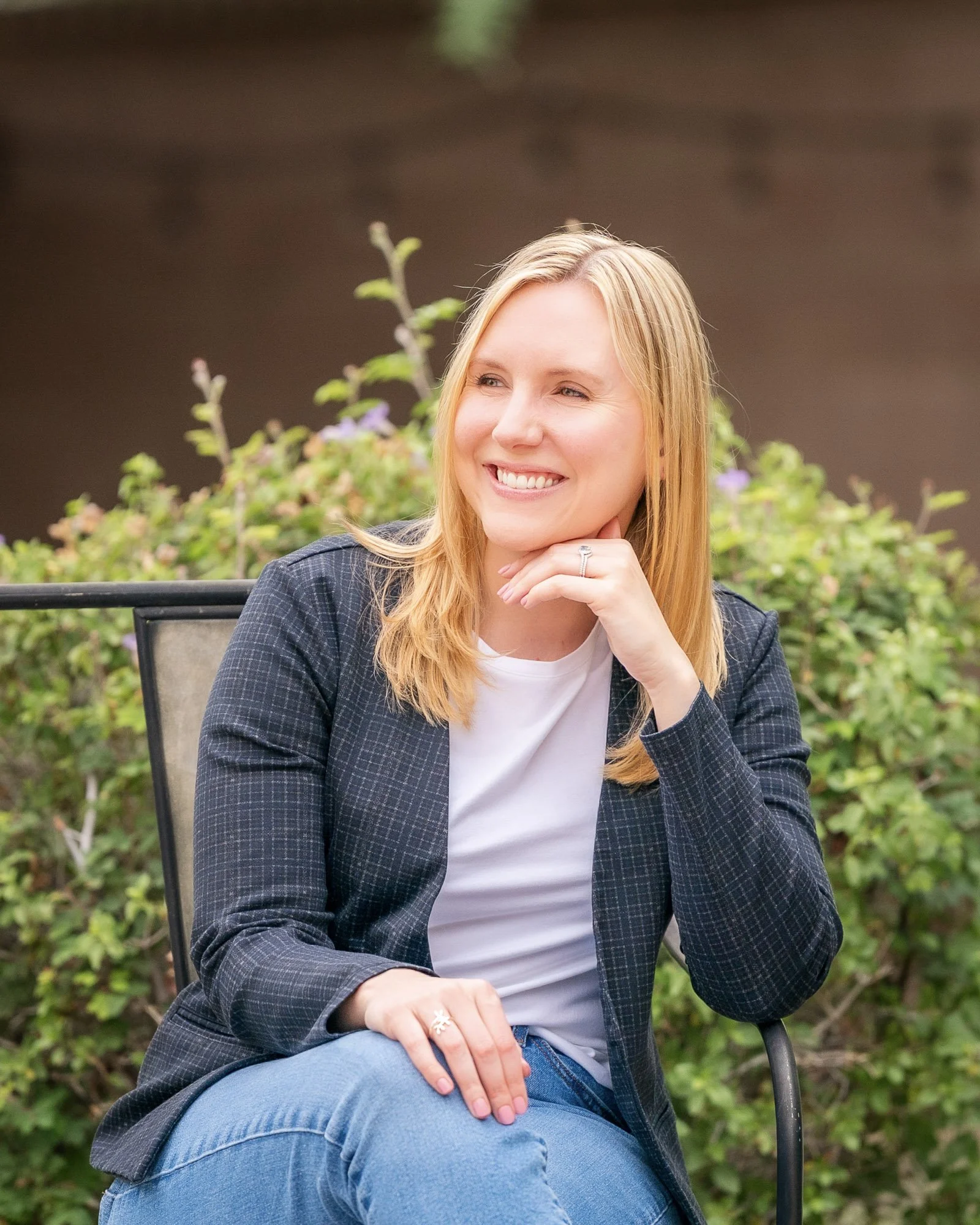 A smiling woman with blonde hair sitting outdoors on a metal chair, wearing a dark blazer, white t-shirt, and blue jeans, with her right hand resting on her chin.