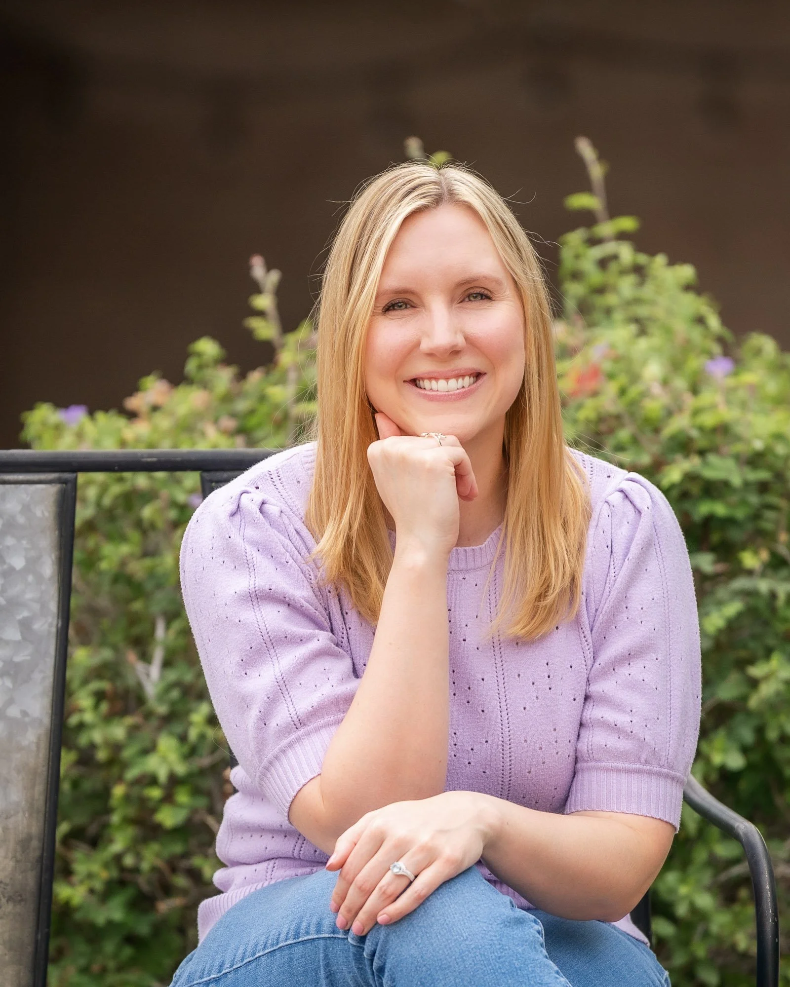 A smiling woman with blonde hair wearing a lavender sweater and blue jeans, sitting outdoors on a bench with greenery in the background.
