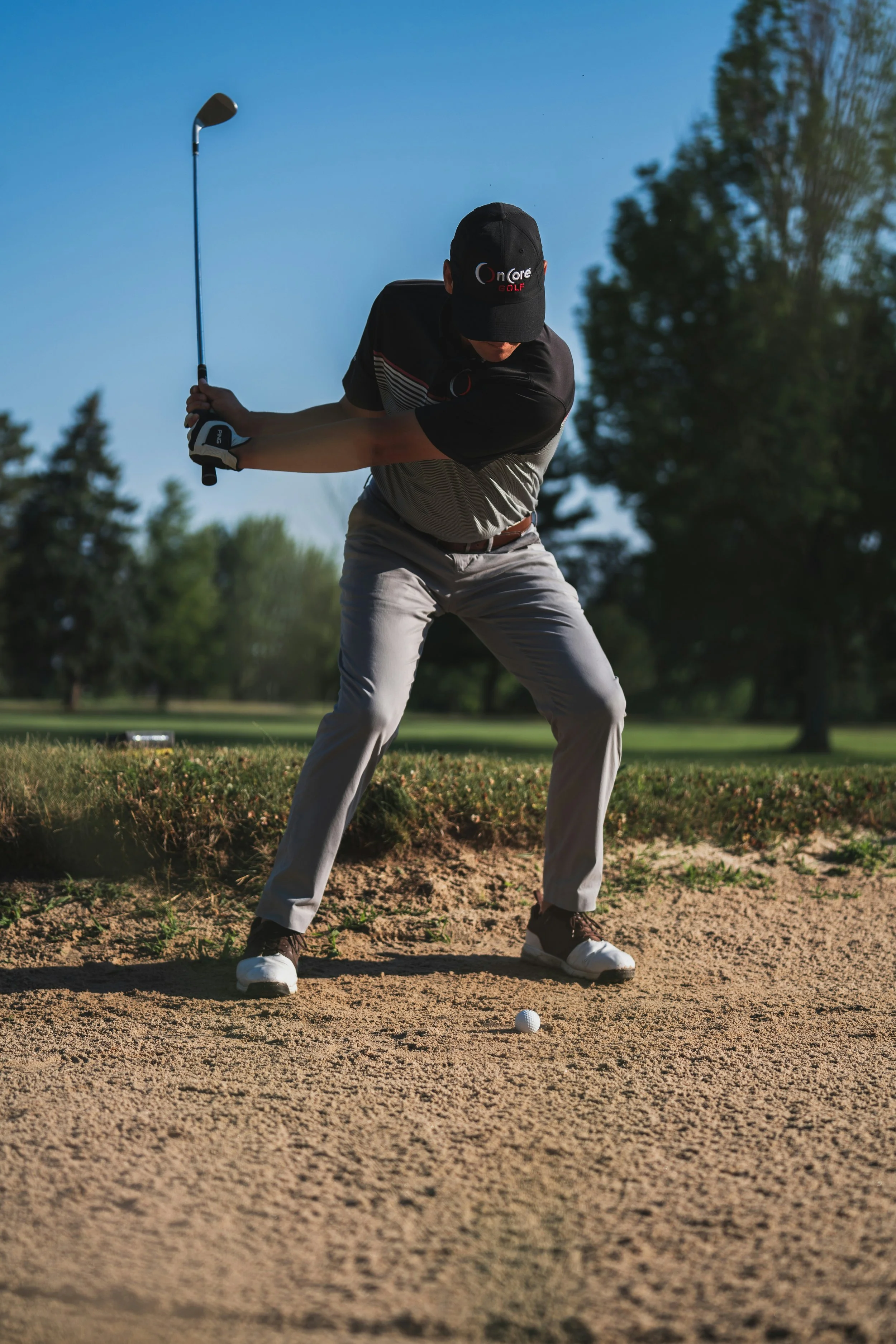 A man in golf attire taking a golf shot from a sand trap on a golf course during daytime.