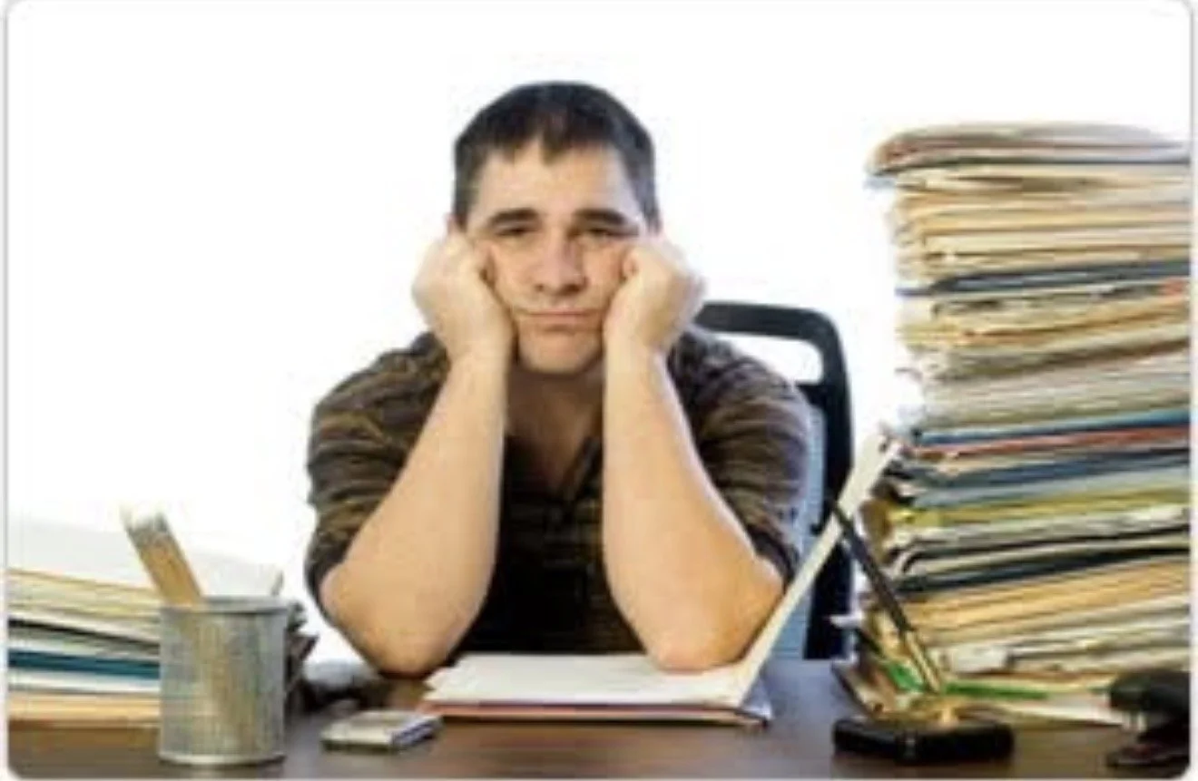 Young man sitting at a cluttered desk, looking bored and tired, with a large stack of papers on the right and office supplies scattered around.