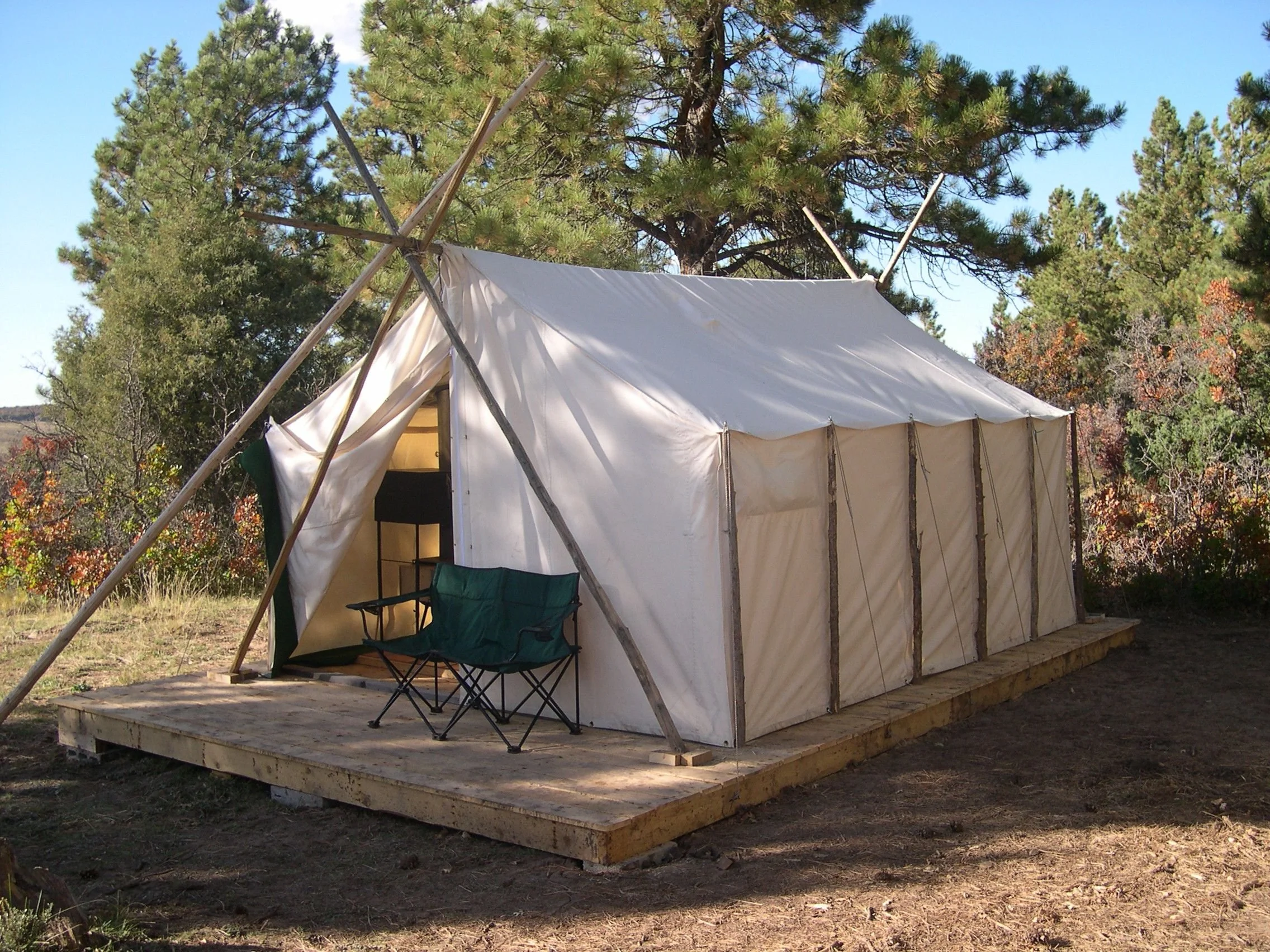 Canvas wall tent set up in a wooded campsite, featuring a peaked roof, straight side walls, and a wood-burning stove chimney.