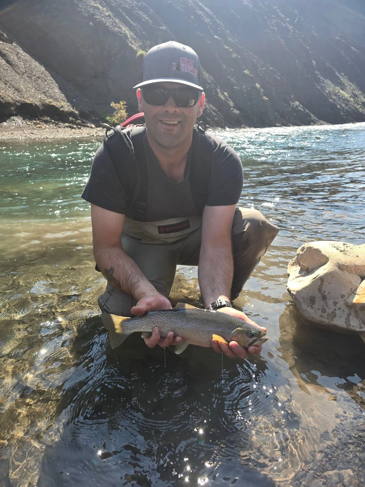 A man wearing sunglasses, a black cap, and a fishing vest kneels in a river, holding a caught rainbow trout with both hands. The river is clear, with rocks visible beneath the surface, and mountainous terrain is in the background.