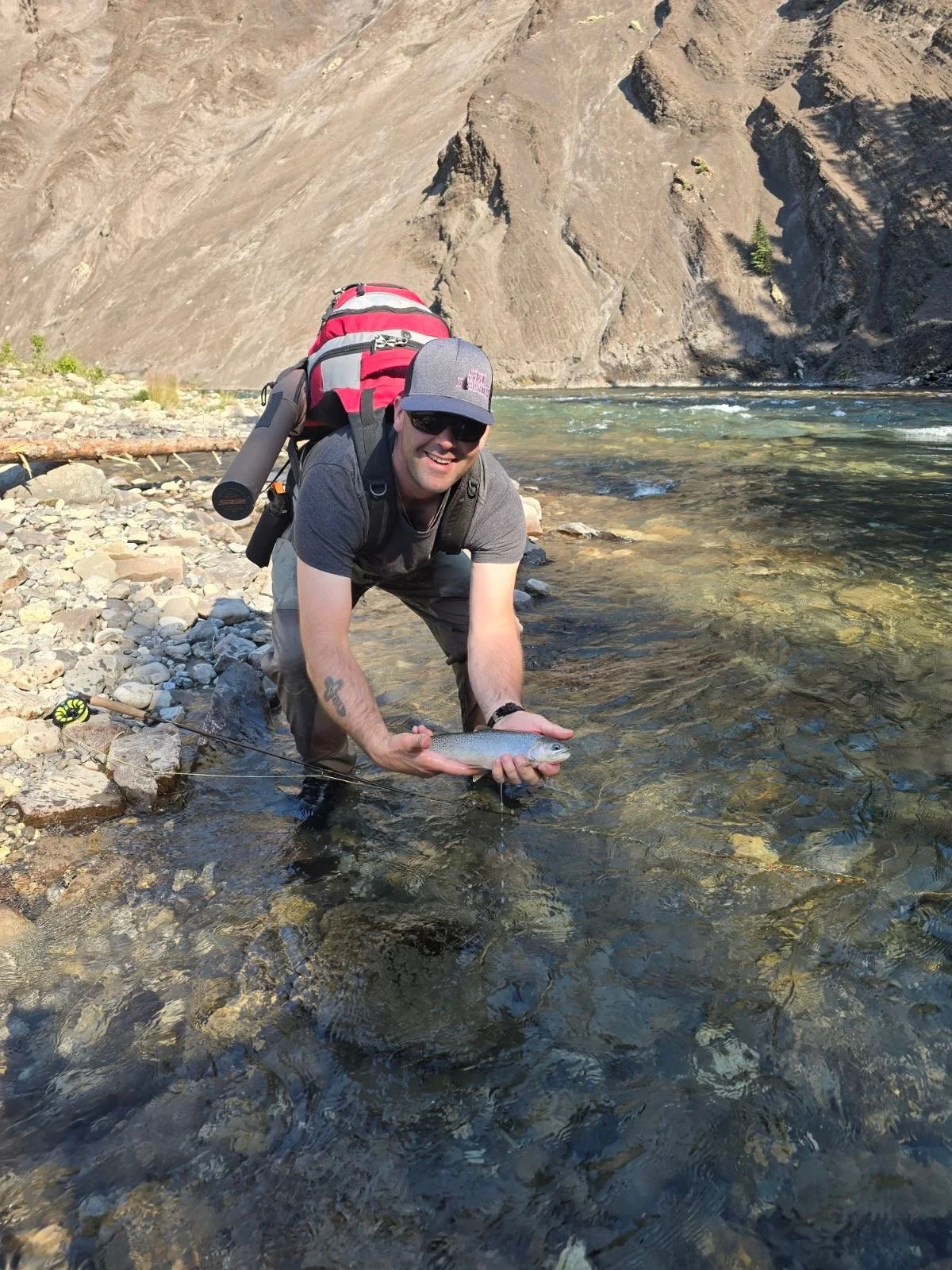 A man wearing sunglasses, a gray cap, and casual outdoor clothing is kneeling in a shallow river, holding a fish with both hands. He has a backpack with gear on his back and is smiling at the camera. The rocky riverbank and tall cliff in the background are visible.