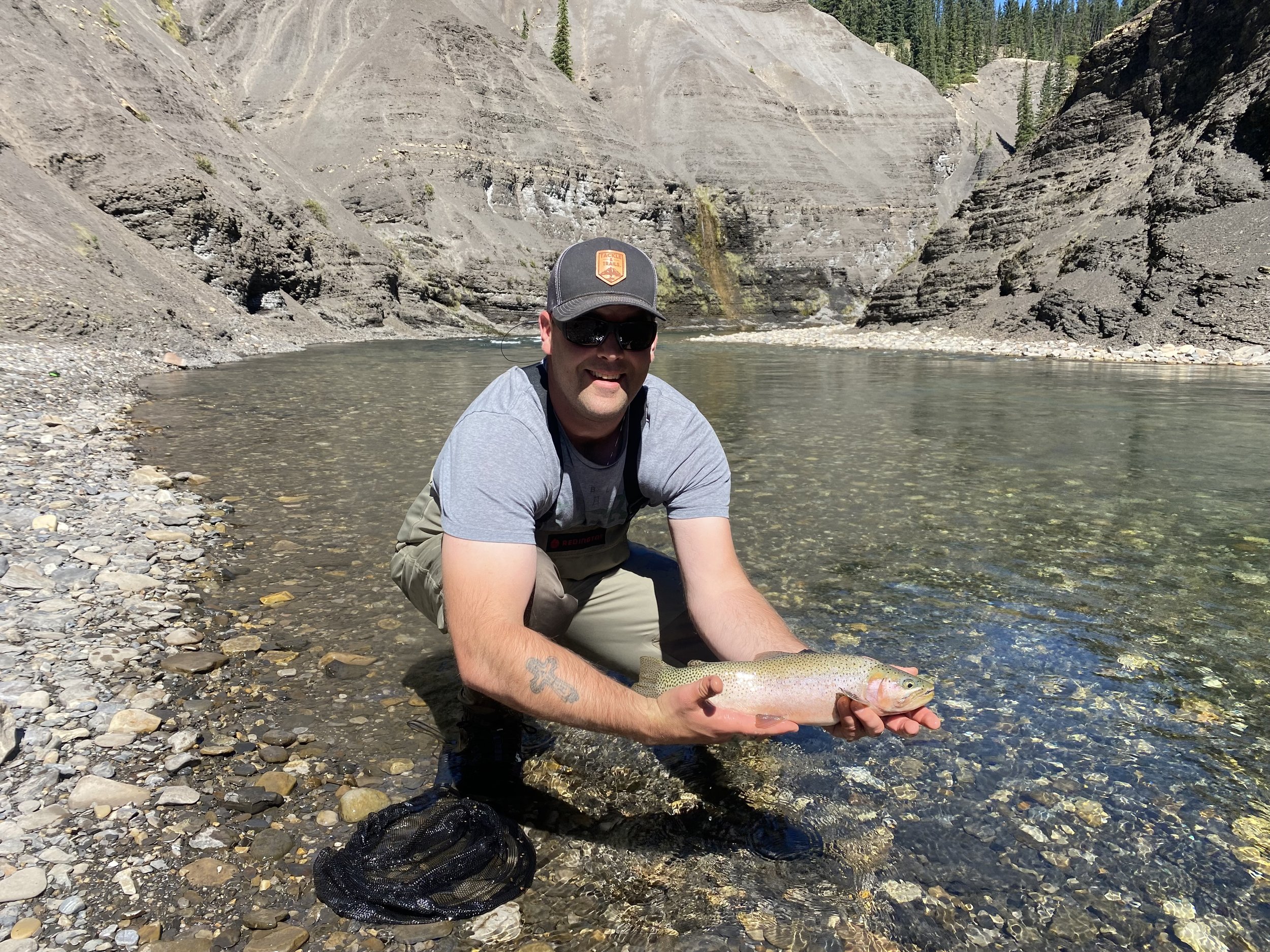 A man wearing sunglasses, a gray t-shirt, and khaki pants kneels by a river holding a large rainbow trout he just caught, with rocky slopes and trees in the background.