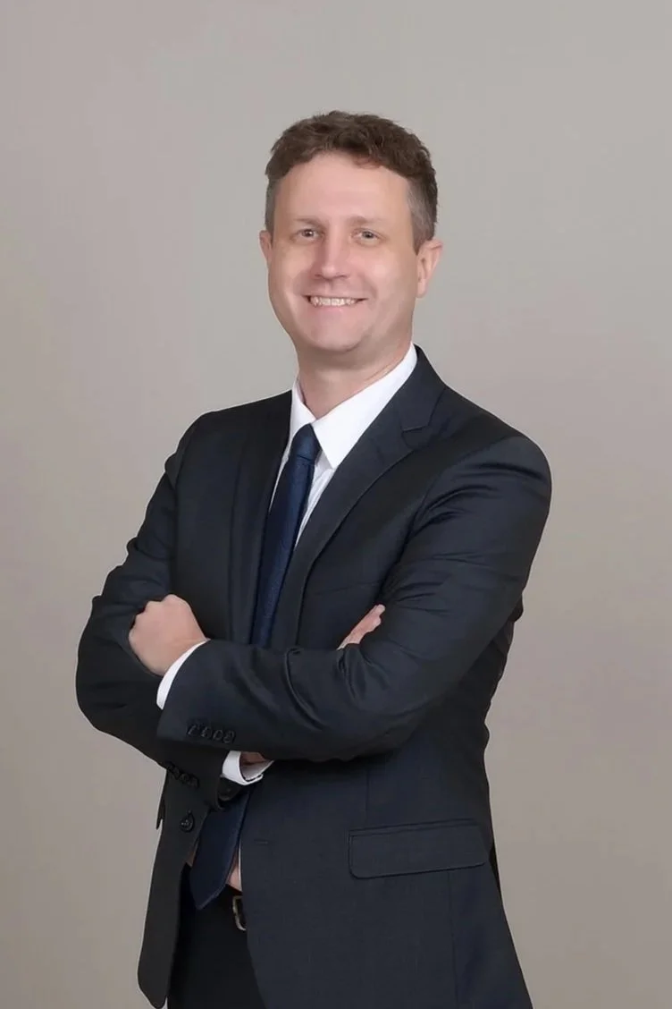 A man in a dark suit and tie smiling with arms crossed in front of a plain background.