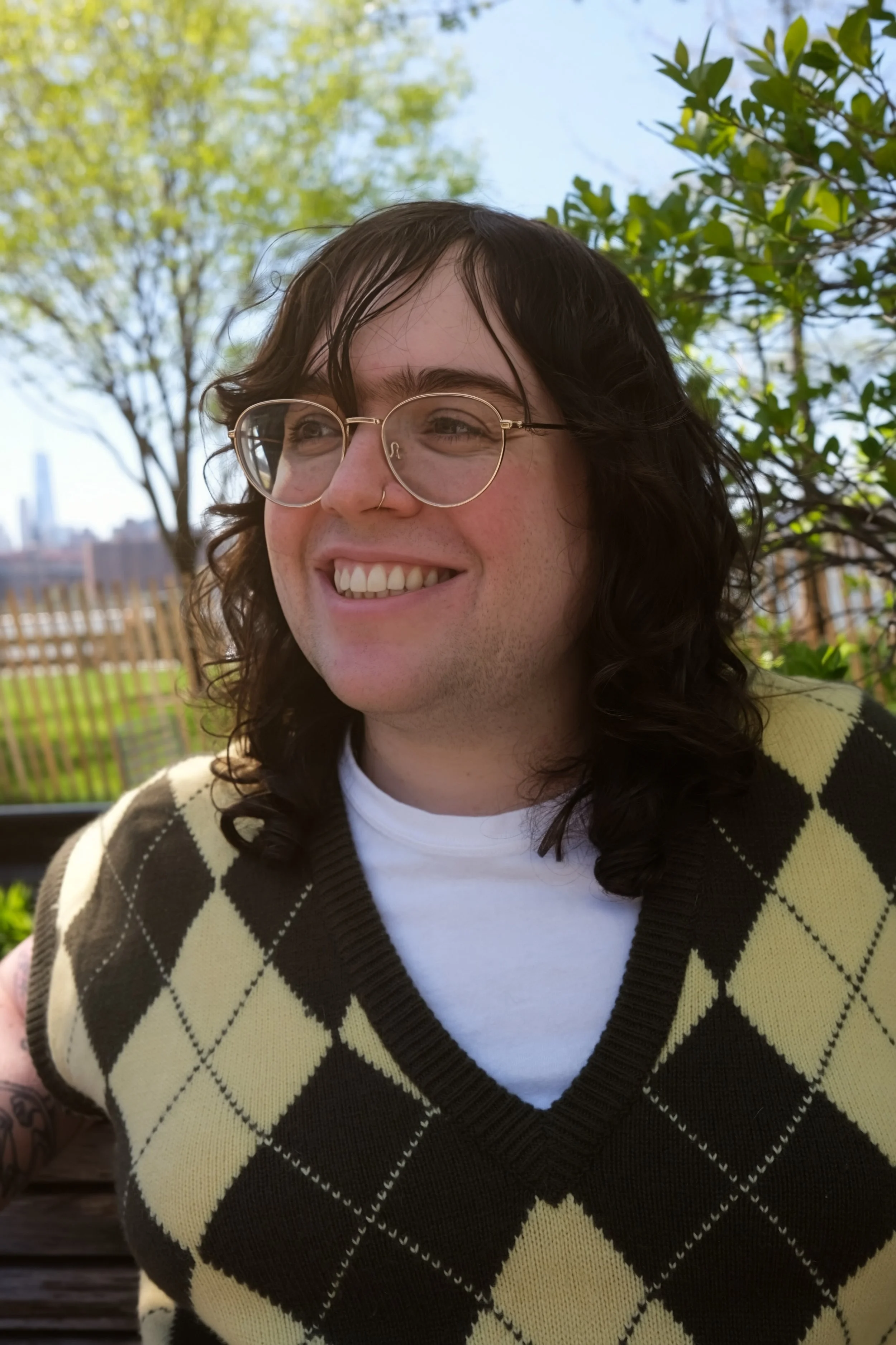 A smiling queer non-binary mental health therapist with long dark hair, glasses, nose ring, wearing a yellow and brown argyle sweater, outdoors in NYC with trees and a fence in the background.