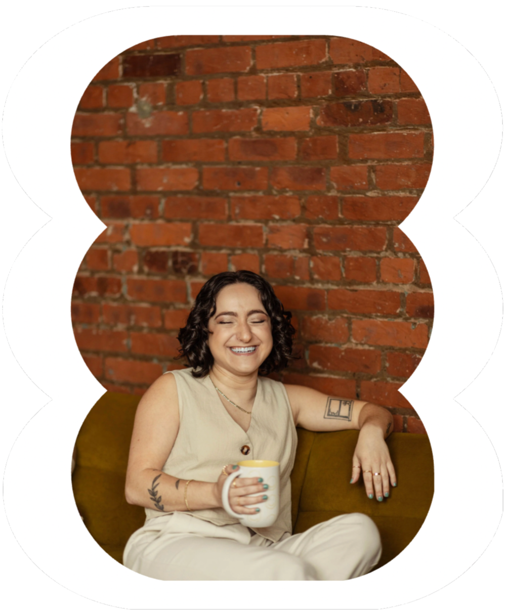 A white queer woman therapist with short curly hair and tattoos on her arm, wearing a beige sleeveless top and beige pants, sitting on a mustard-colored couch with a brick wall background, holding a white mug and smiling with eyes closed.