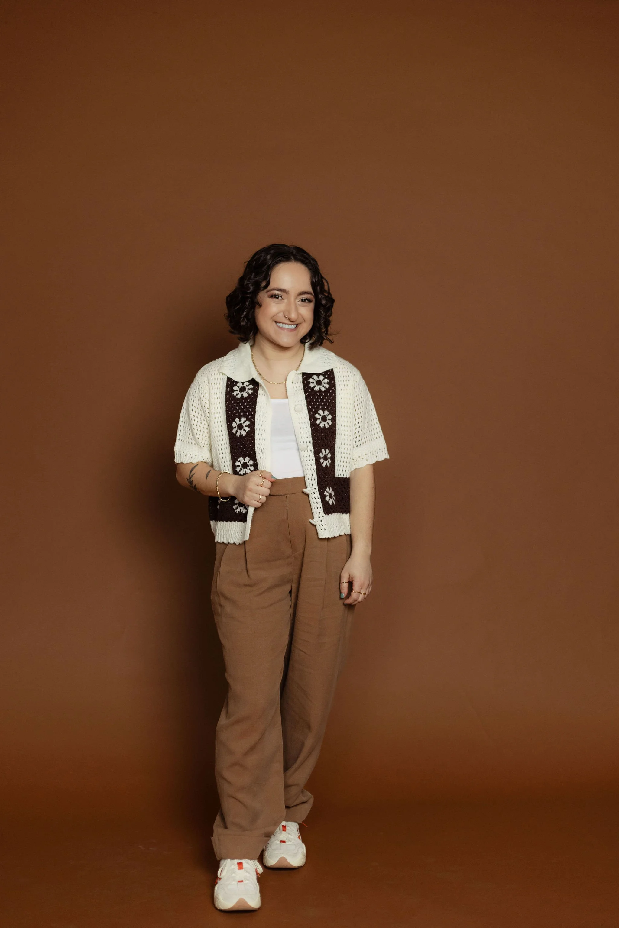A female LGBTQ+ mental health therapist with short curly hair smiling, wearing a white t-shirt, a cream and brown patterned flower knit cardigan, brown high-waisted pants, and white sneakers, standing against a solid brown background.