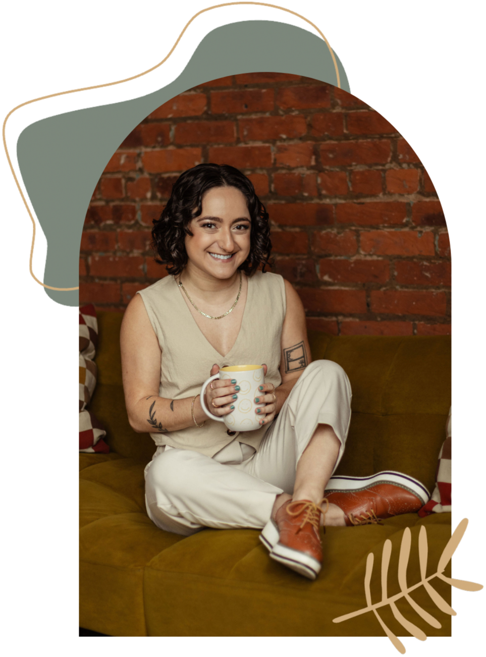 A queer woman mental health therapist with short dark curly hair sitting on a yellow therapy sofa, holding a yellow and white mug, smiling at the camera, with a brick wall in the background. She has tattoos on her arms and is wearing a beige vest