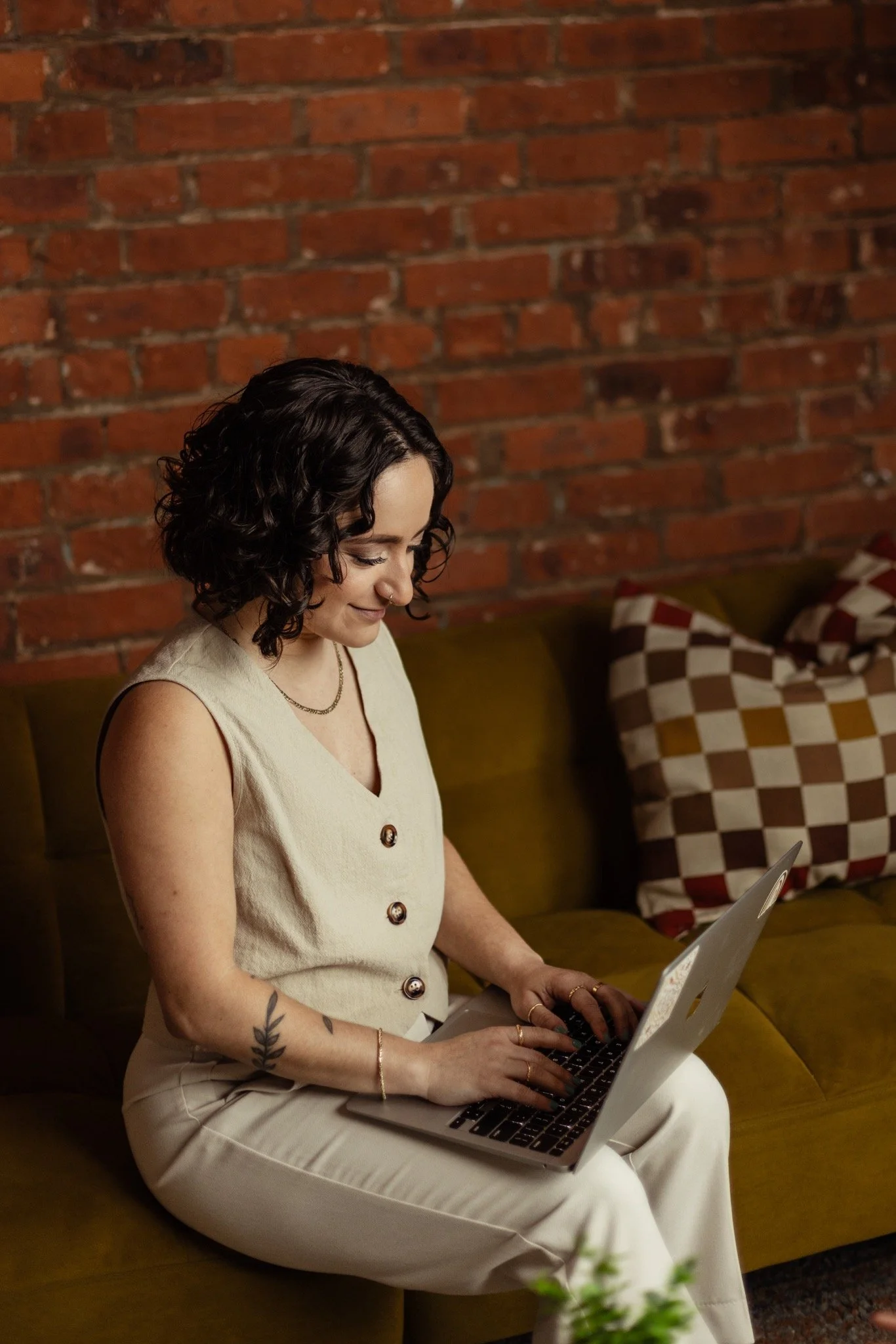 A white queer woman therapist with short curly hair and tattoos on her arm, wearing a beige sleeveless top and beige pants, sitting on a yellow velvet couch, using a laptop, in front of a brick wall and checkered patterned pillows.