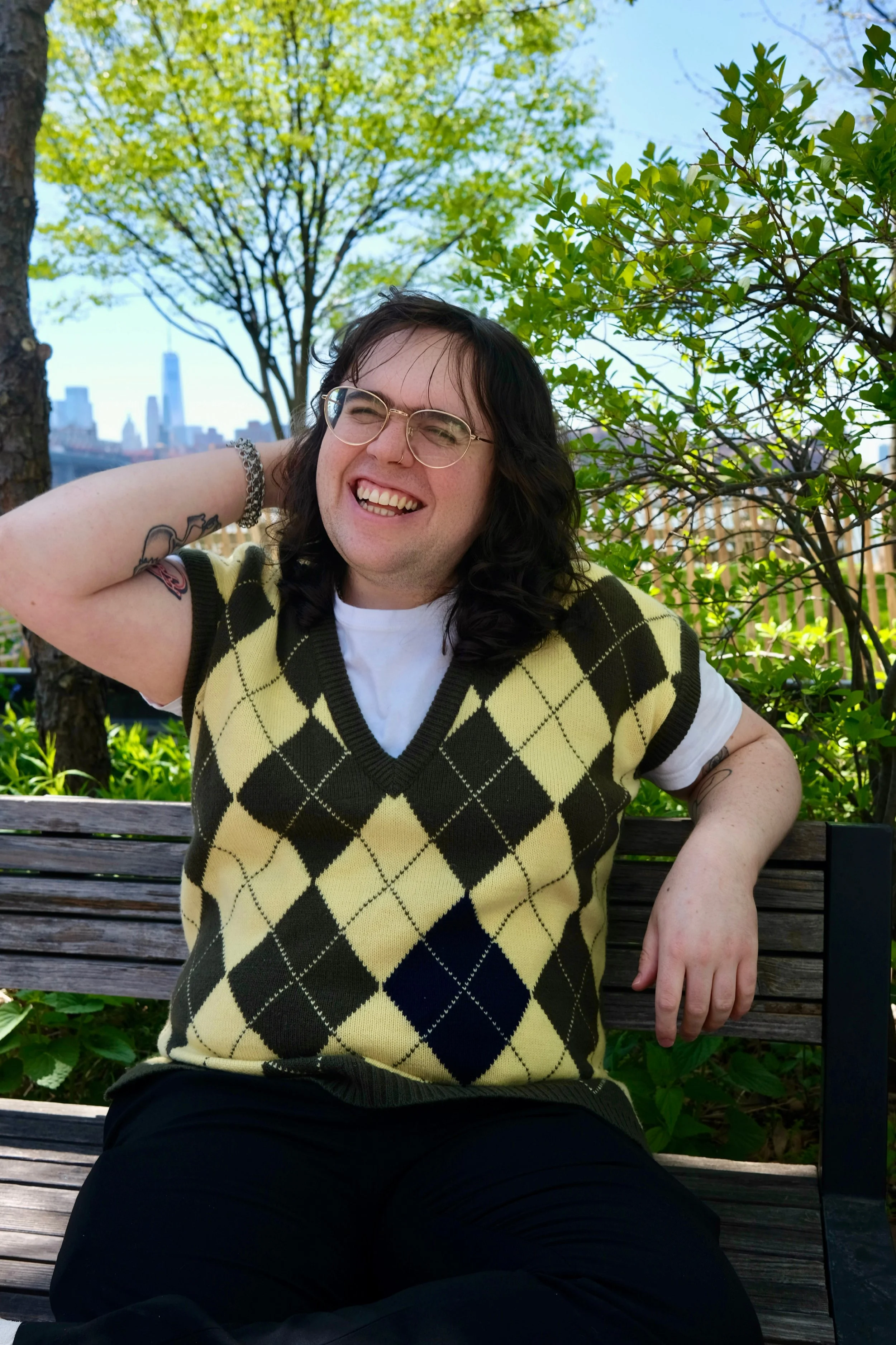A transfemme mental health therapist with curly brown hair wearing glasses, a yellow and brown argyle sweater vest, sitting on a bench outdoors. Smiling widely with arm behind head, looking off to she side. Has tattoos and a bracelet on her left arm.