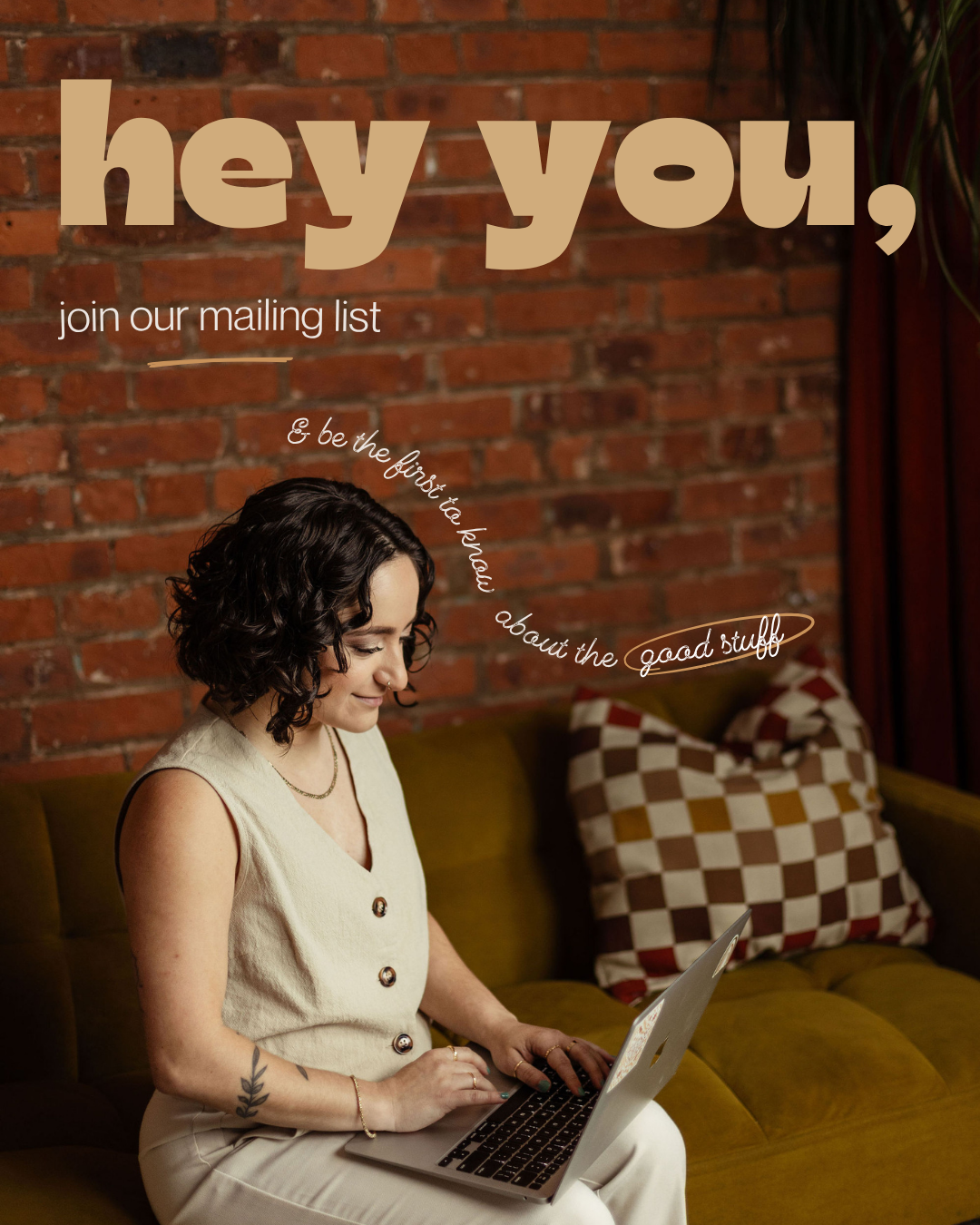 A smiling queer woman mental health therapist with short curly dark hair sitting on a vintage yellow sofa with a brick wall background, using a laptop, with text overlay inviting to join a mailing list for free mental health resources and updates.