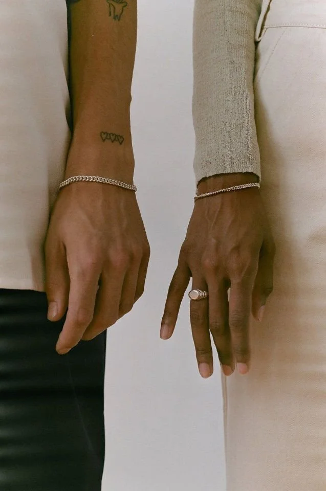 Close-up of two hands and wrists, each wearing a bracelet; one hand has a ring, and both have tattoos and are positioned next to each other.