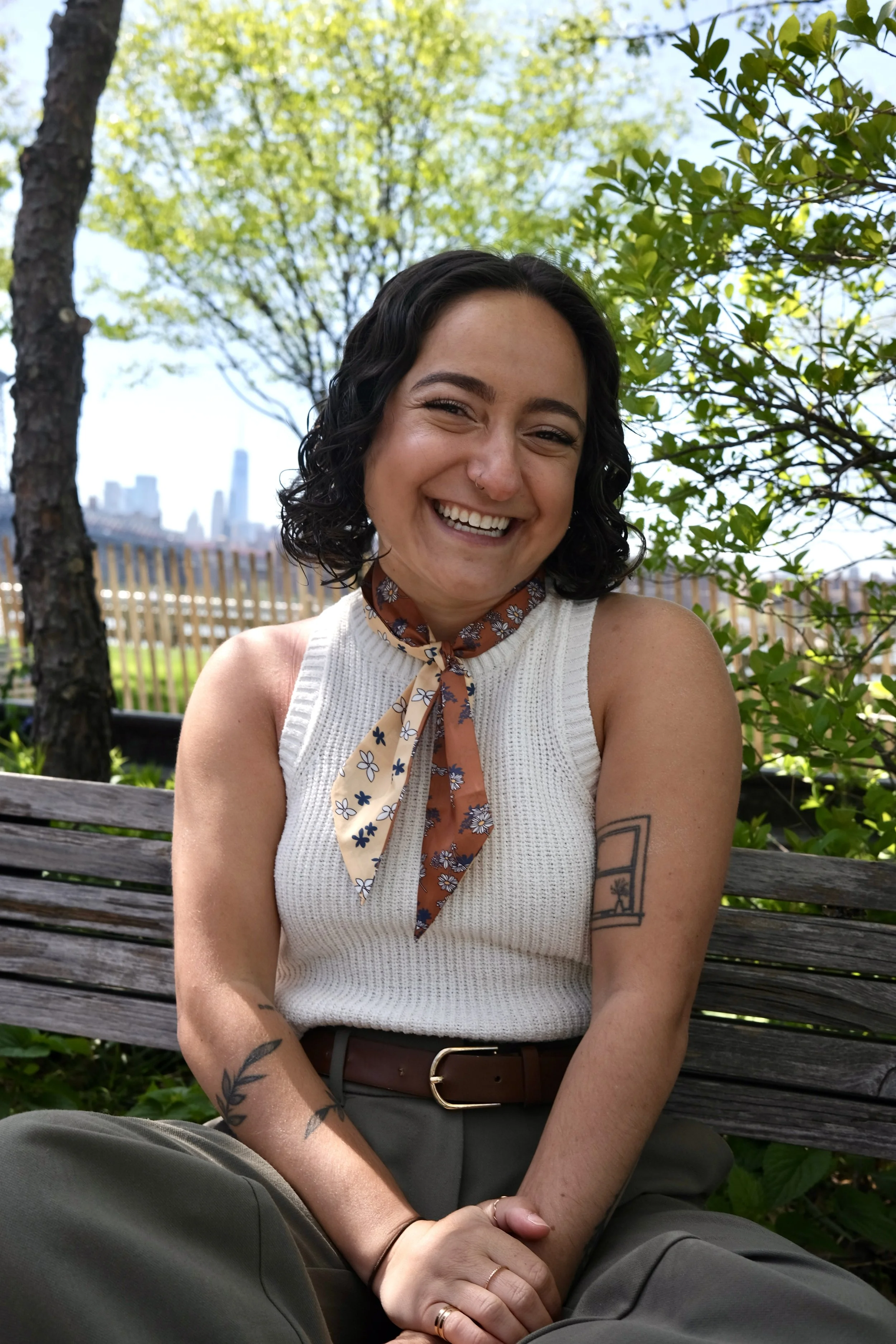 A smiling queer woman mental health therapist with short curly dark hair outdoors on a wooden bench, with trees and a city skyline in the background. She has tattoos on her arms and is wearing a white sleeveless top, a floral scarf, and green pants.