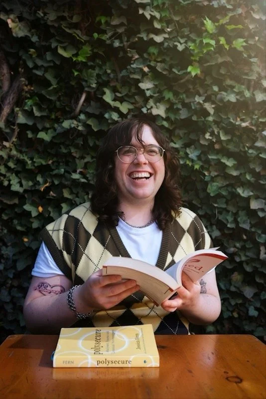 A non-binary mental health therapist with glasses and tattoos  sitting at a wooden table outdoors, smiling and reading a book. There is a second book titled 'Polysecure' on the table in front of them, with a background of green ivy covering a wall.