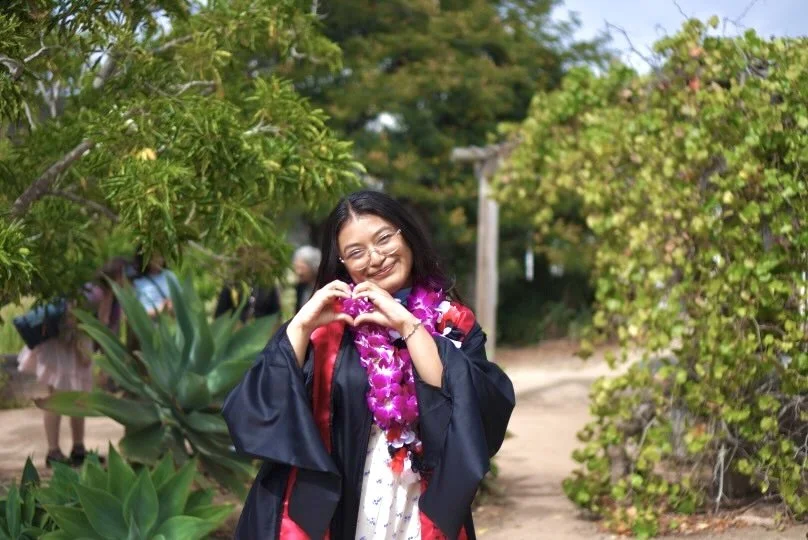 A woman in a graduation gown and lei making a heart shape with her hands, smiling outdoors with greenery around her.