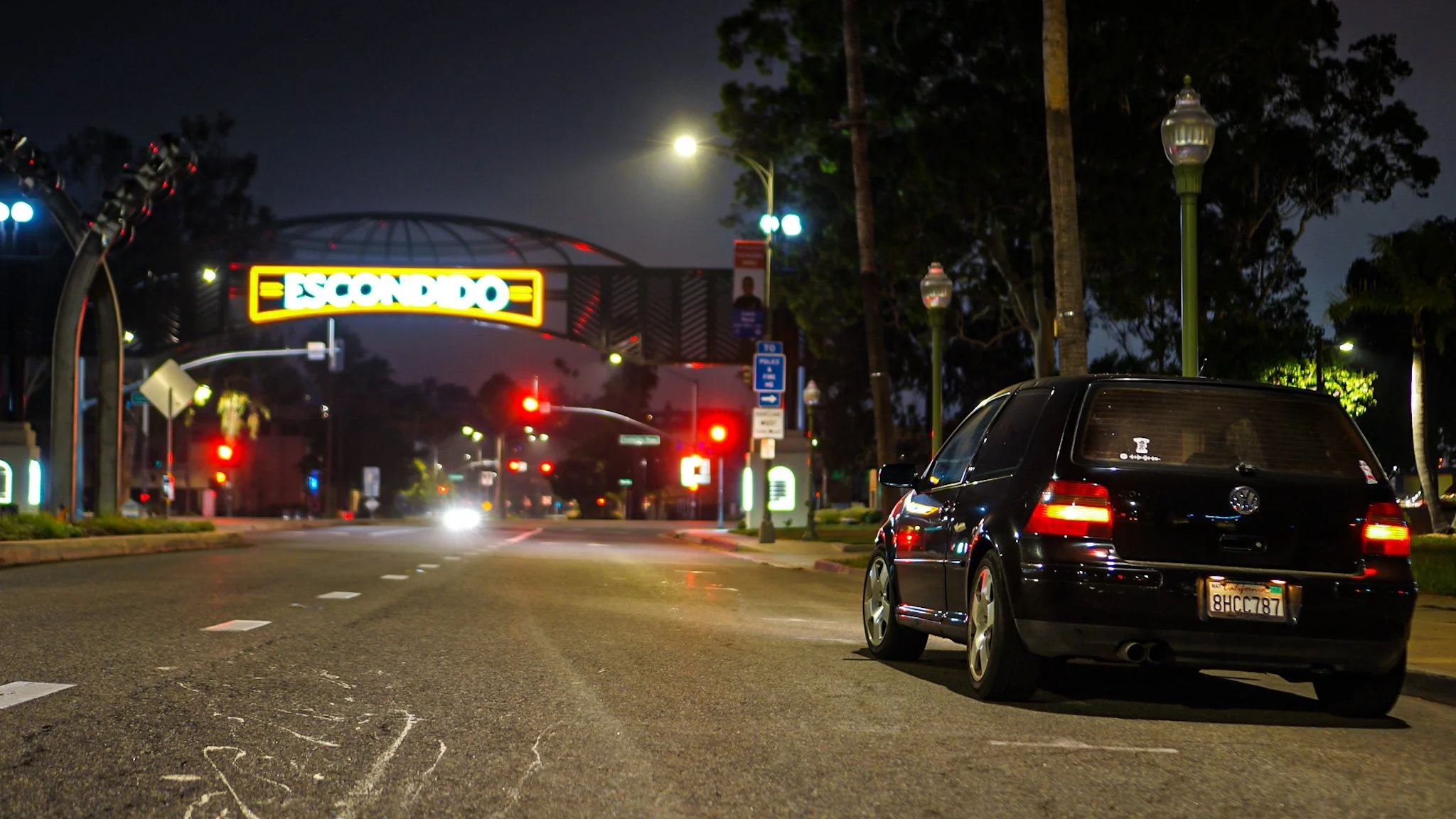A dark street scene at night with a black Volkswagen Golf parked on the right side, illuminated streetlights, a large digital sign with the word "ESCONDIDO" in the distance, and traffic lights with red signals. Trees line the sidewalk on the right, and the sky is cloudy or overcast.
