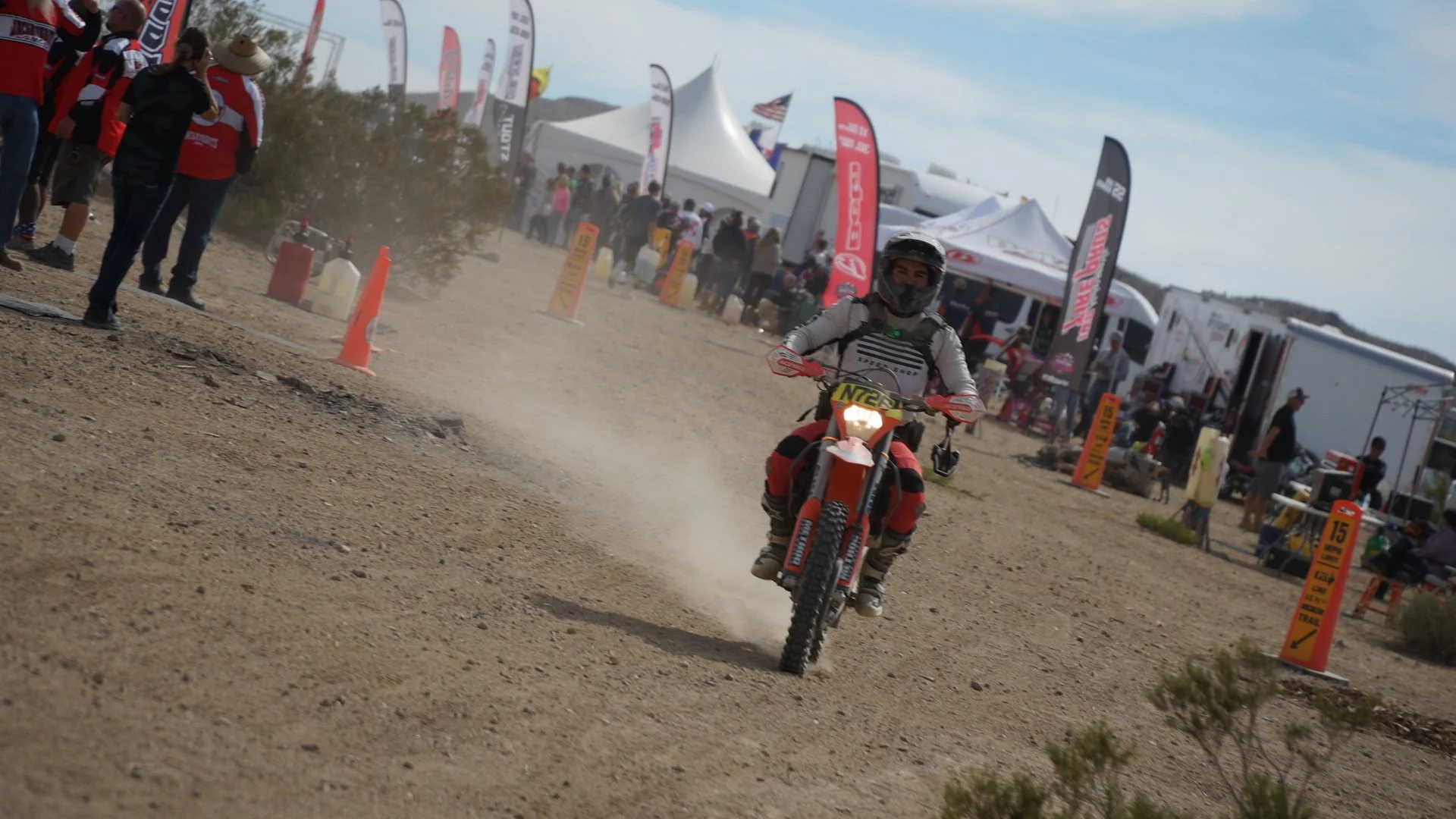A person riding a dirt bike on a dusty track at an outdoor event, with tents, banners, and spectators in the background.