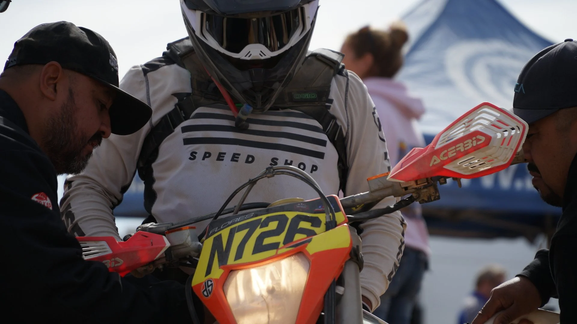 Motocross rider with helmet and gear being helped by two crew members before a race, with a blue tent and a woman in the background.