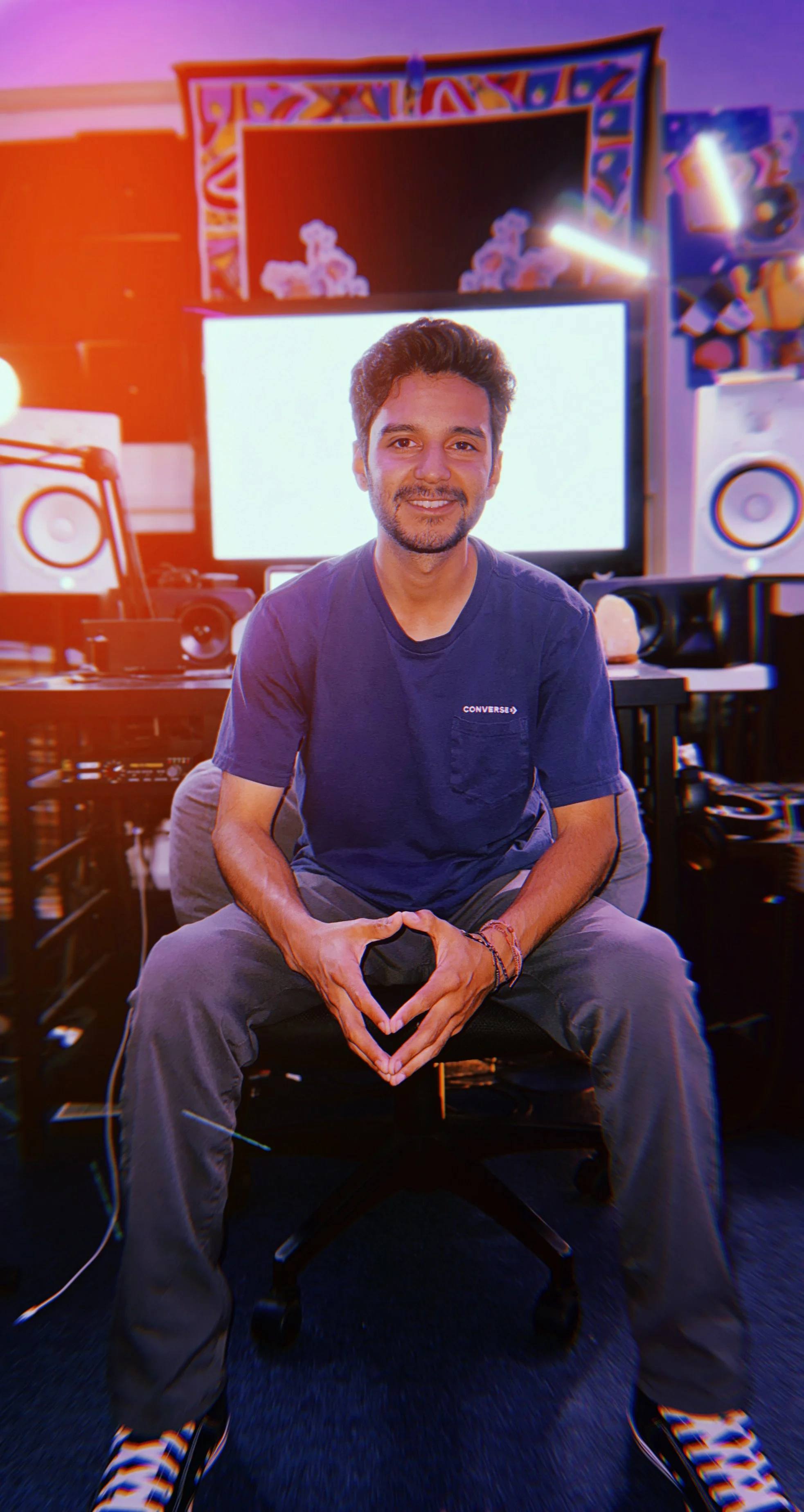 A man sitting in a music studio with electronic music equipment, speakers, and a large monitor behind him, smiling at the camera.