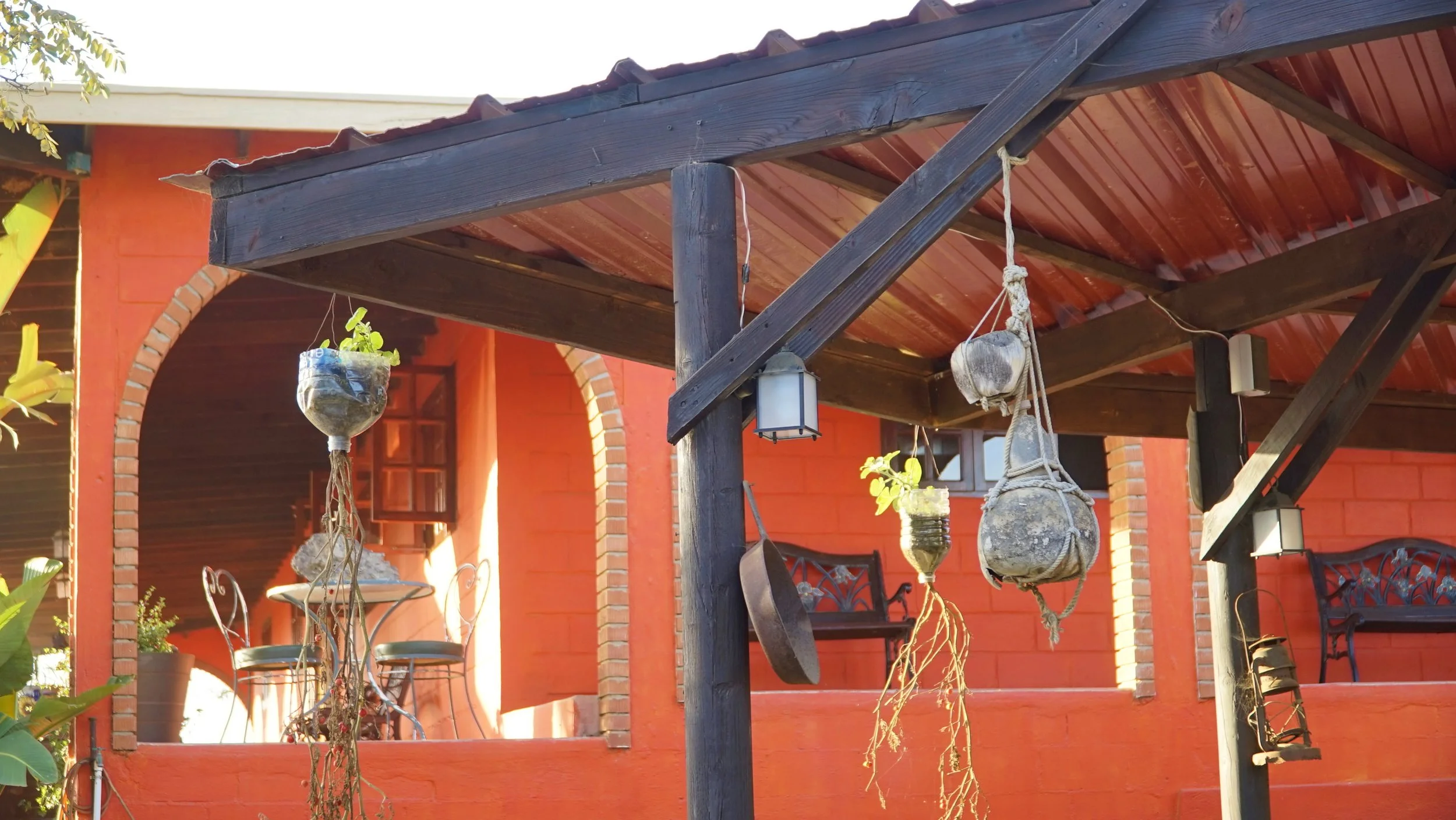 Courtyard with hanging plant pots, a hanging lantern, and a hat, with a red building featuring brick arches and outdoor furniture in the background.