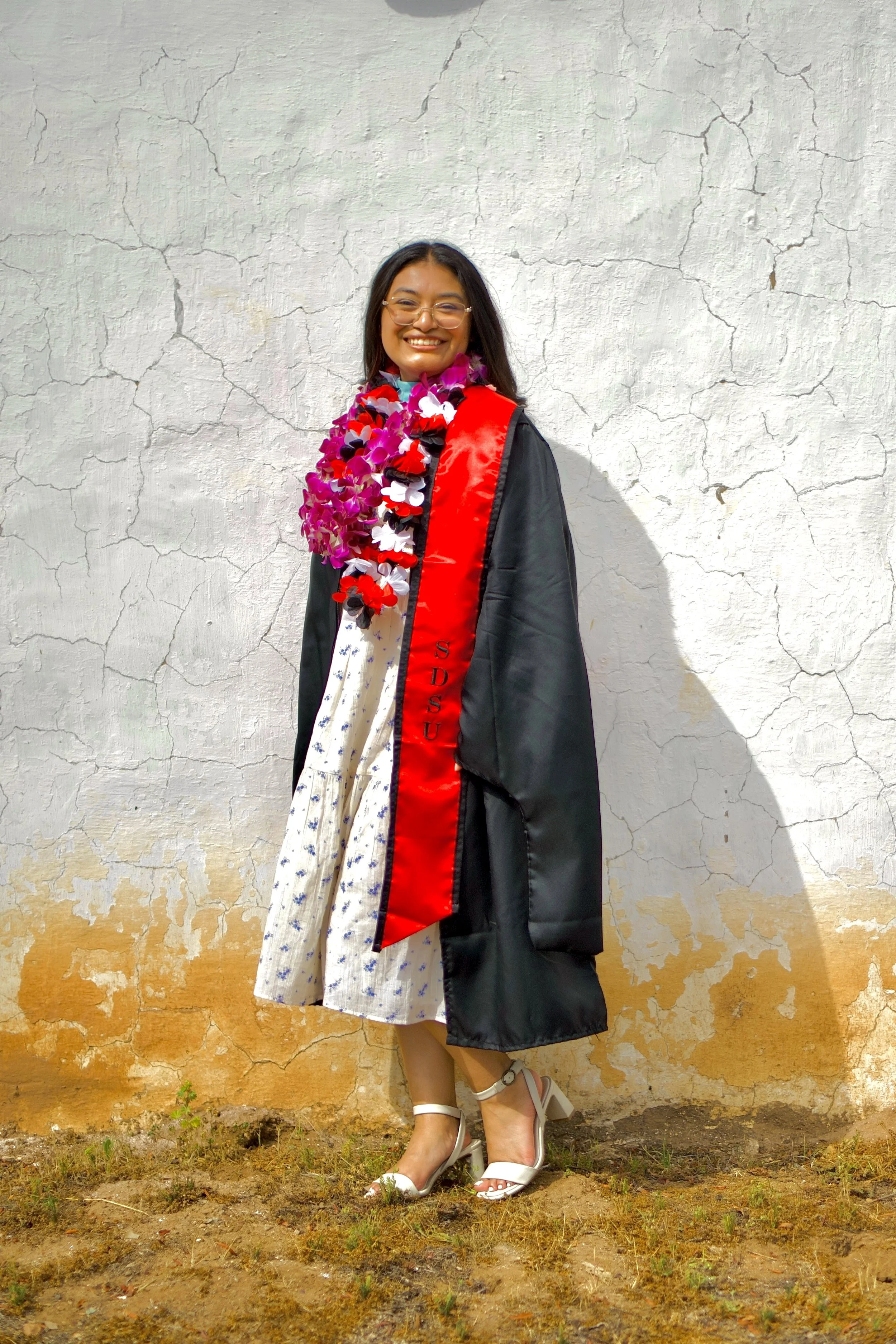 A woman in graduation attire wearing a cap and gown, with a red and black stole, standing against a textured white wall, smiling, with a colorful lei around her neck.