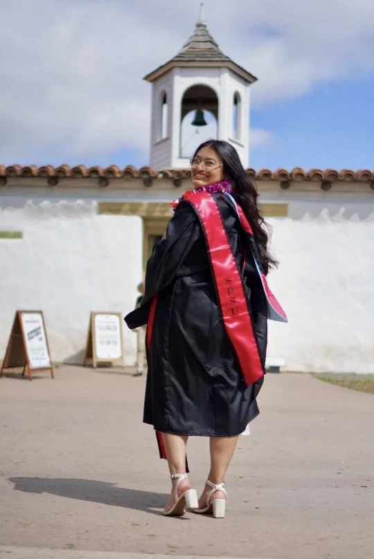 A young woman in a graduation gown and cap, smiling and looking over her shoulder, standing outside in front of a white building with a bell tower, during daytime.